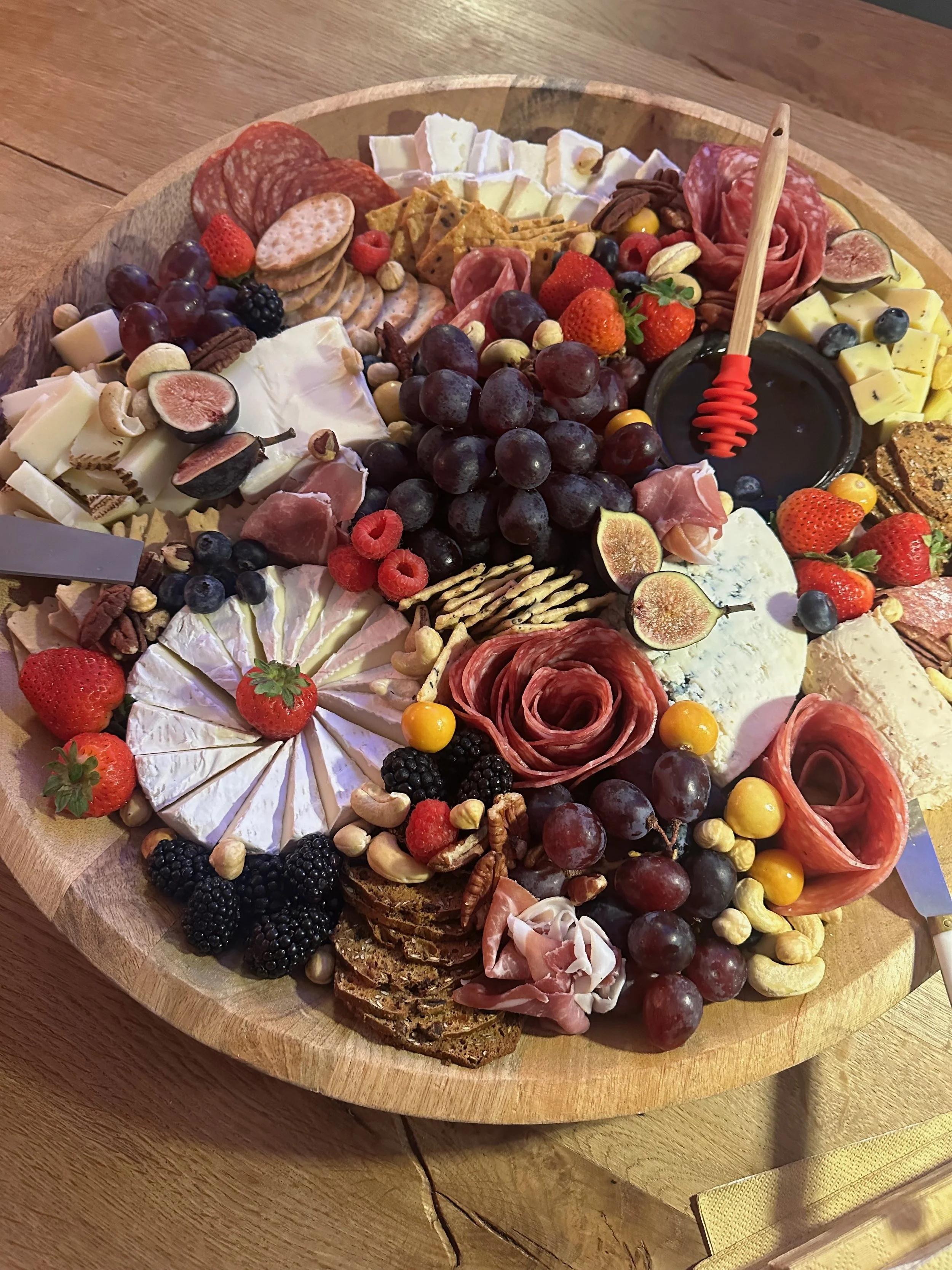A round wooden charcuterie board filled with assorted cheeses, cured meats, fresh berries, crackers, nuts, and fruit slices.