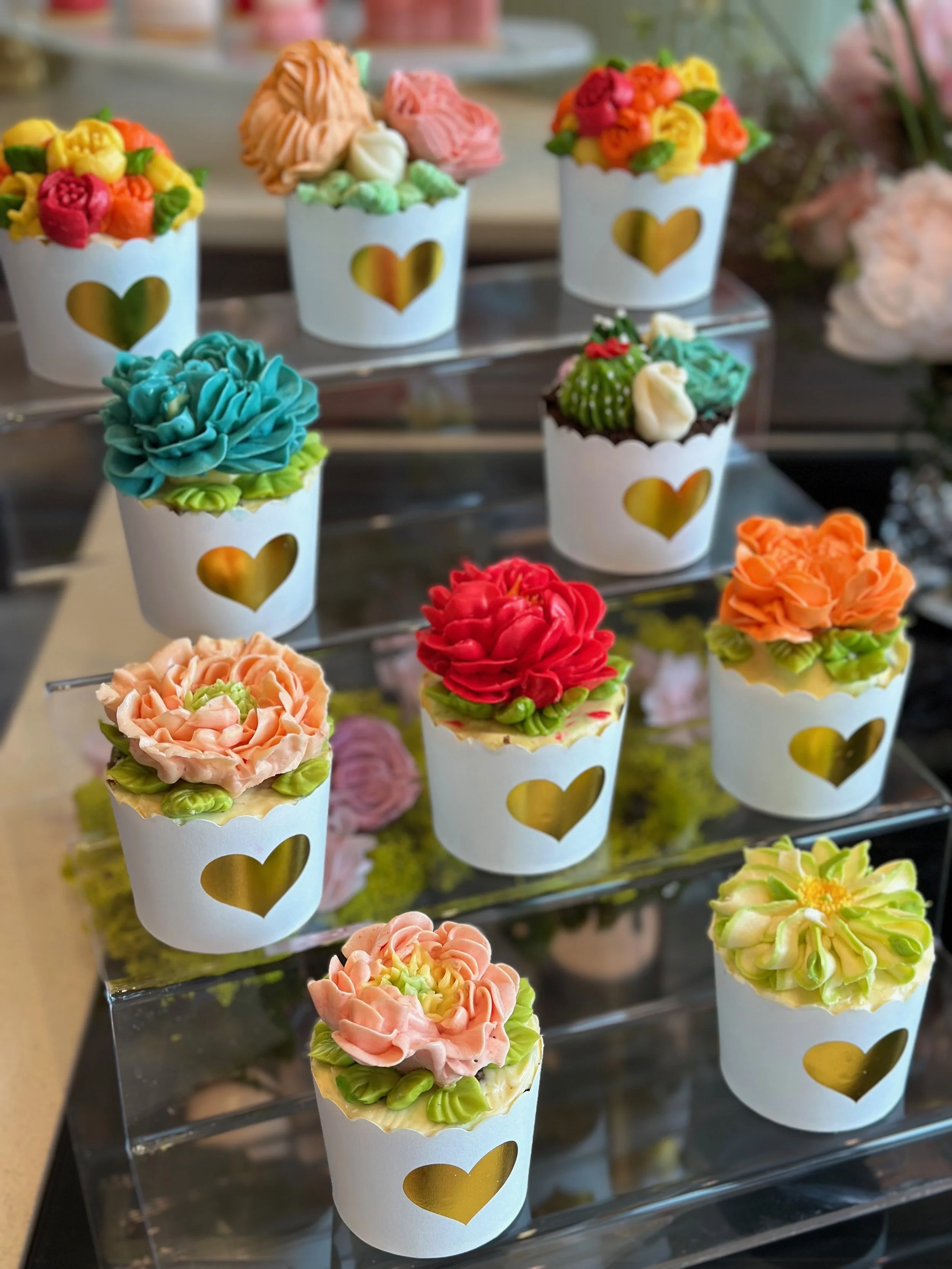 Colorful cupcakes decorated with intricately piped buttercream flowers in various colors, displayed on a tiered clear stand.