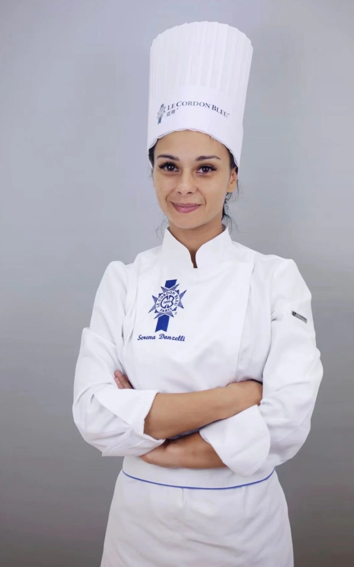 A female chef wearing a white chef's coat with blue logos and a tall white chef's hat, standing with her arms crossed against a gray background.