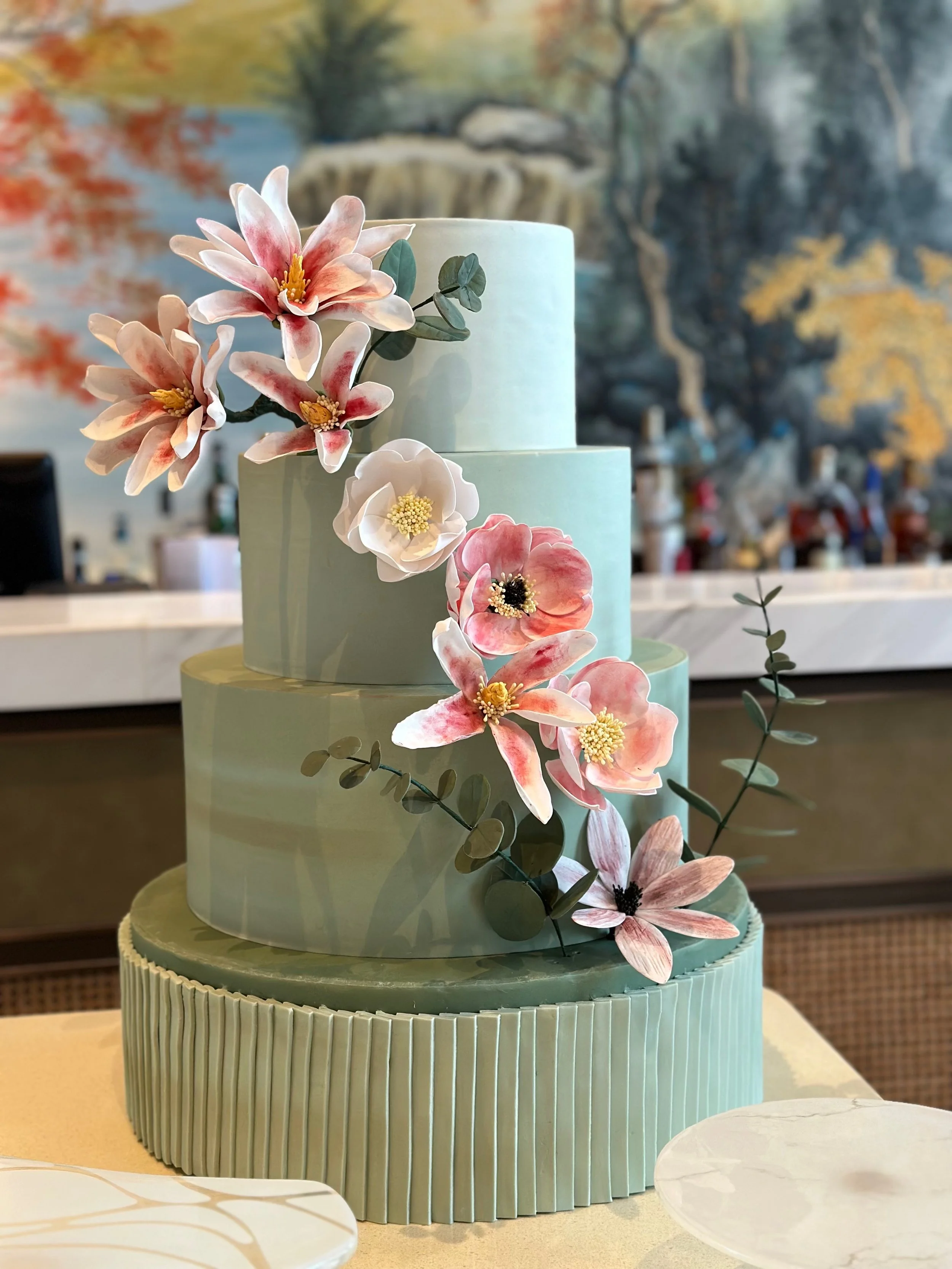 A tiered cake with pastel green fondant, decorated with pink and white flowers and green leaves, displayed on a table.