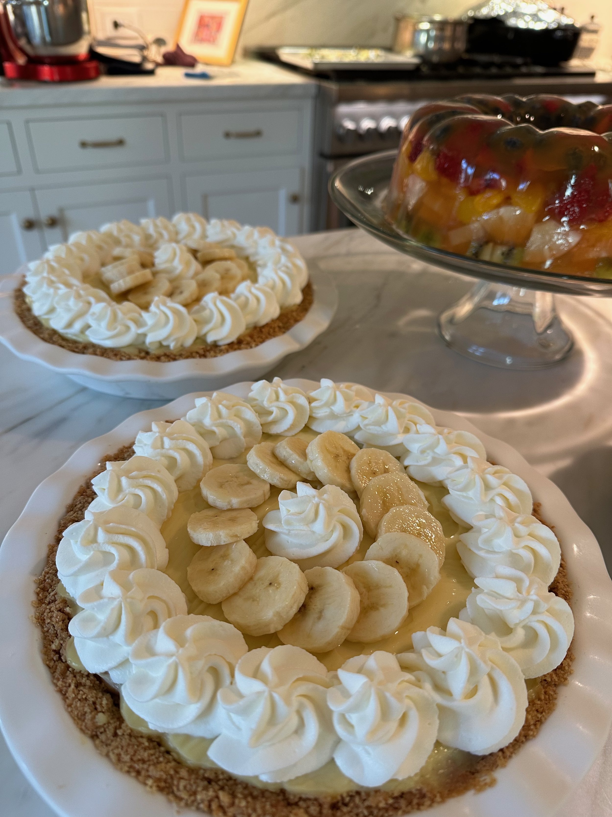 Two banana cream pies topped with sliced bananas and piped whipped cream, with a fruit salad in a glass bowl in the background.