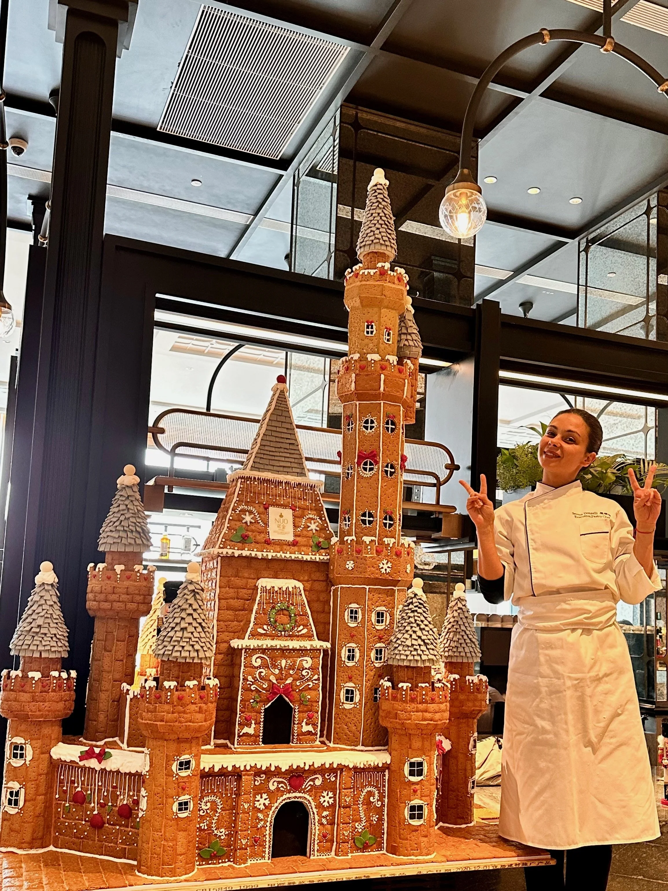 A woman in a white chef's coat standing next to an elaborate gingerbread castle decorated with icing, candies, and gingerbread details, set indoors with modern ceiling and lighting.