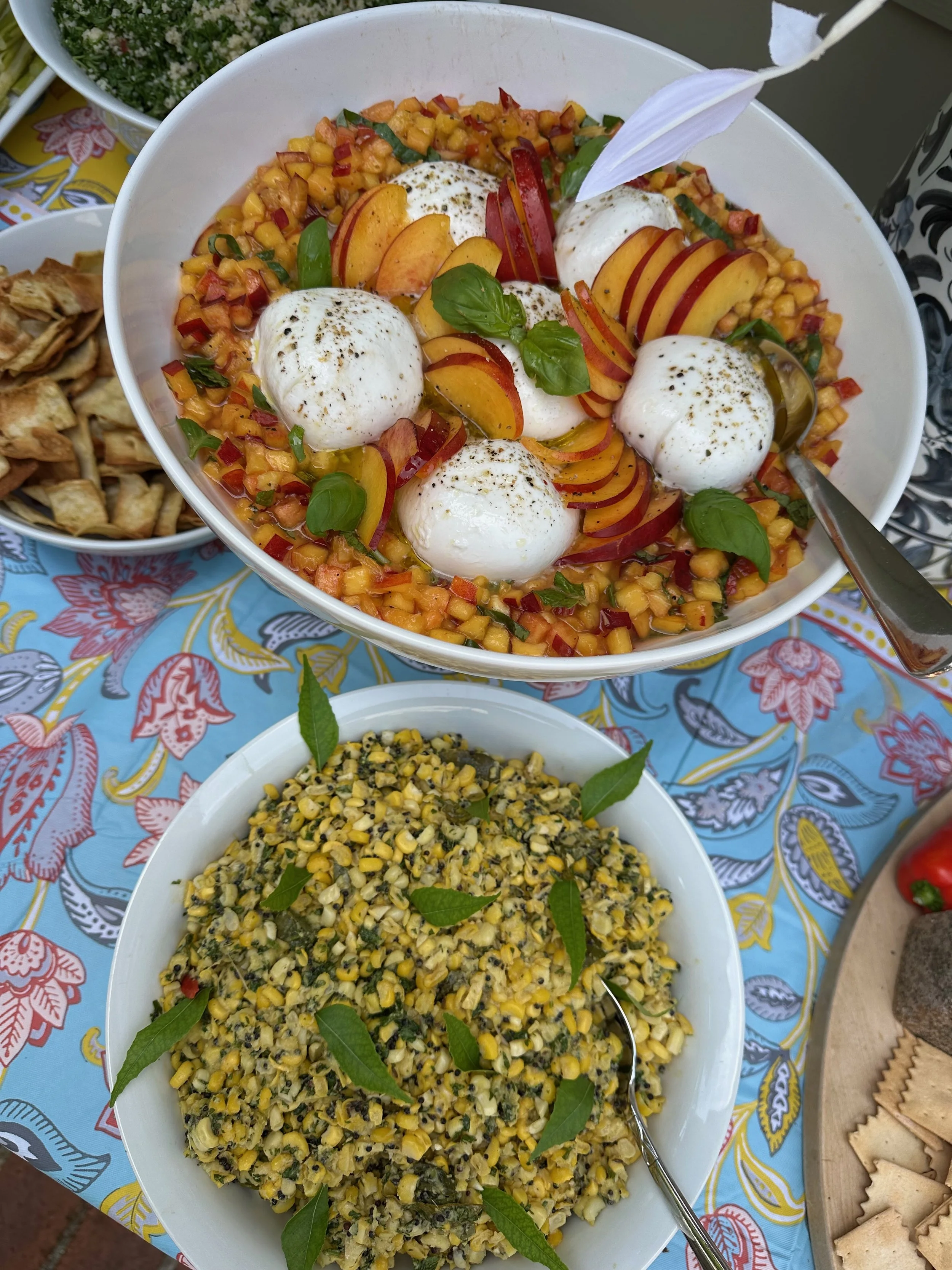 A salad with burrata cheese, peach slices, cherry tomatoes, and fresh basil, served in a white bowl. There is a bowl of corn salad with herbs and a small bowl of roasted vegetables on a colorful tablecloth.