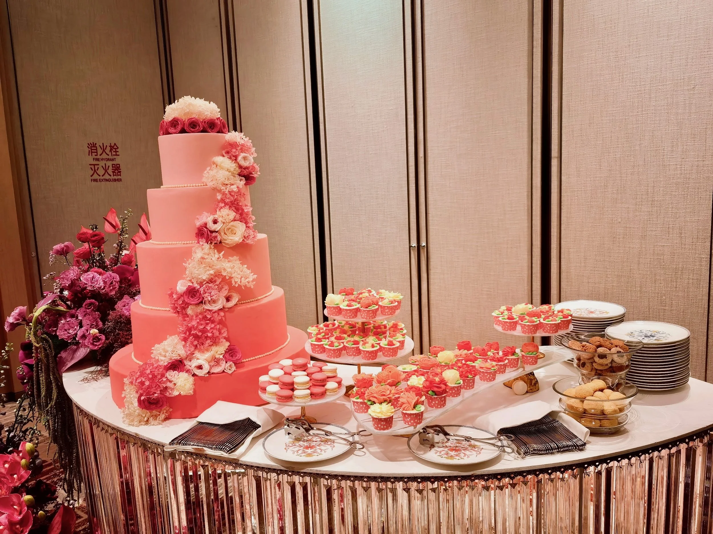 A pink, four-tiered wedding cake decorated with pink and white flowers, surrounded by cupcakes, cookies, and other desserts on a dessert table.