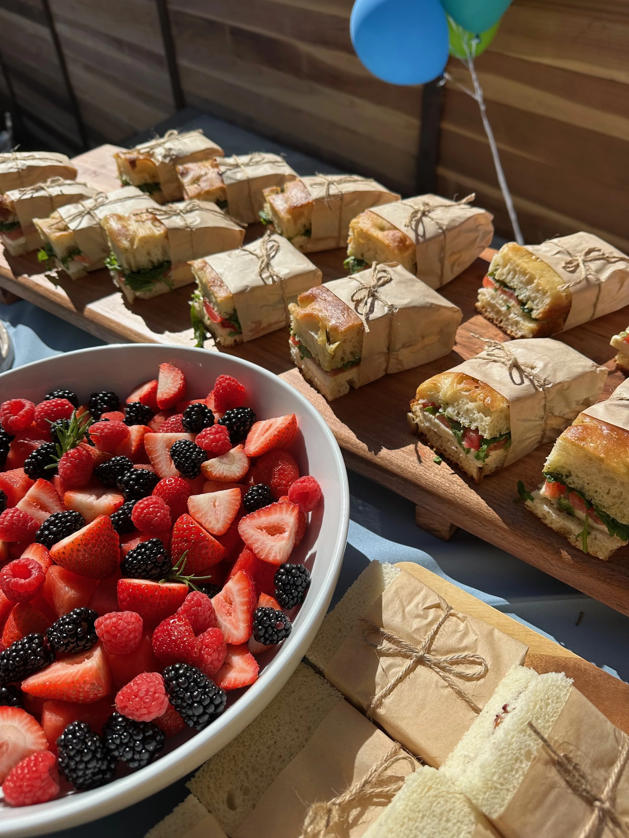 A platter of mixed berries including strawberries, raspberries, blackberries, and red and black raspberries next to a tray of sandwiches wrapped in brown paper and tied with string, on a table with balloons in the background.