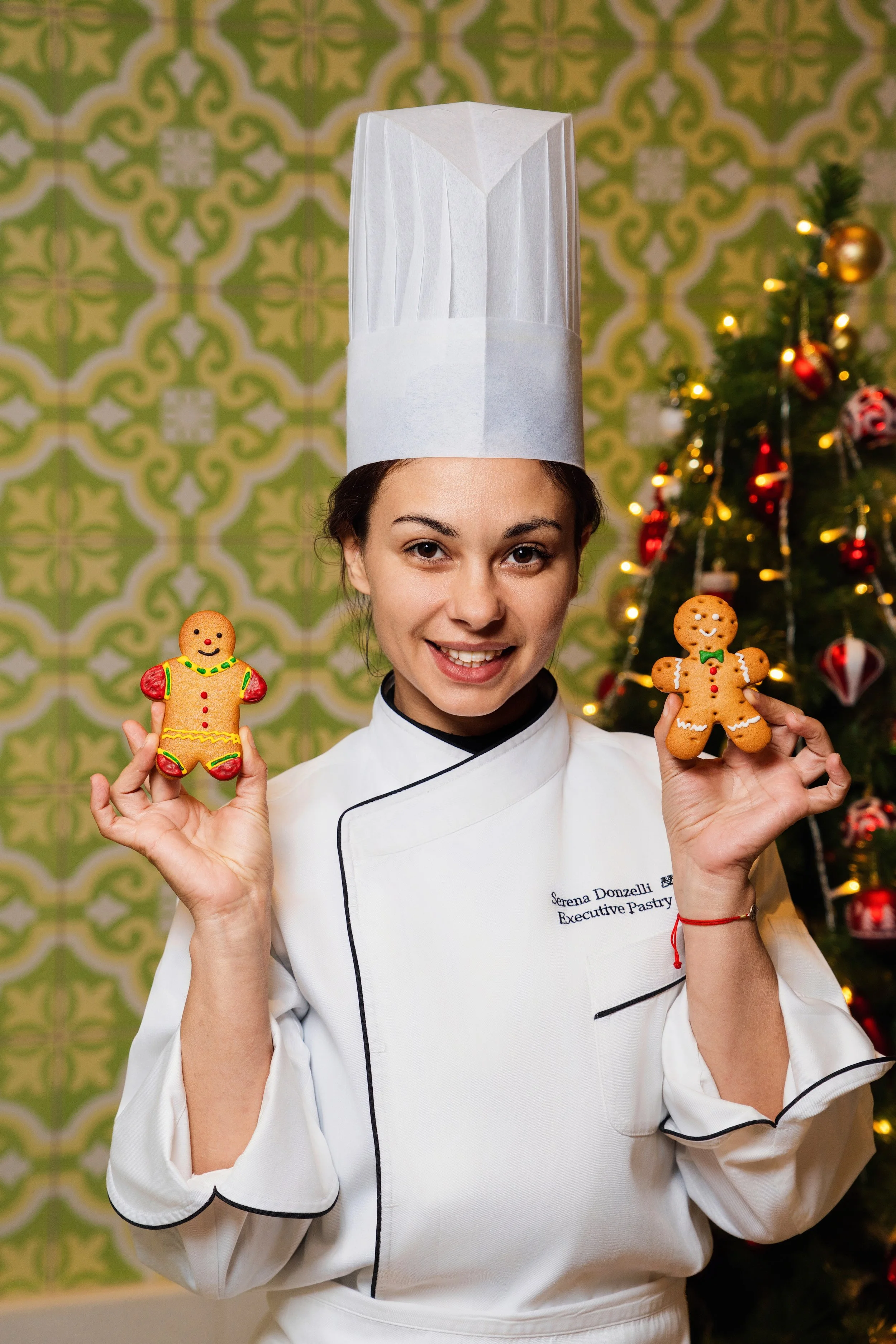 A female chef in a white uniform and chef hat holding decorated gingerbread cookies, smiling, with a Christmas tree in the background.