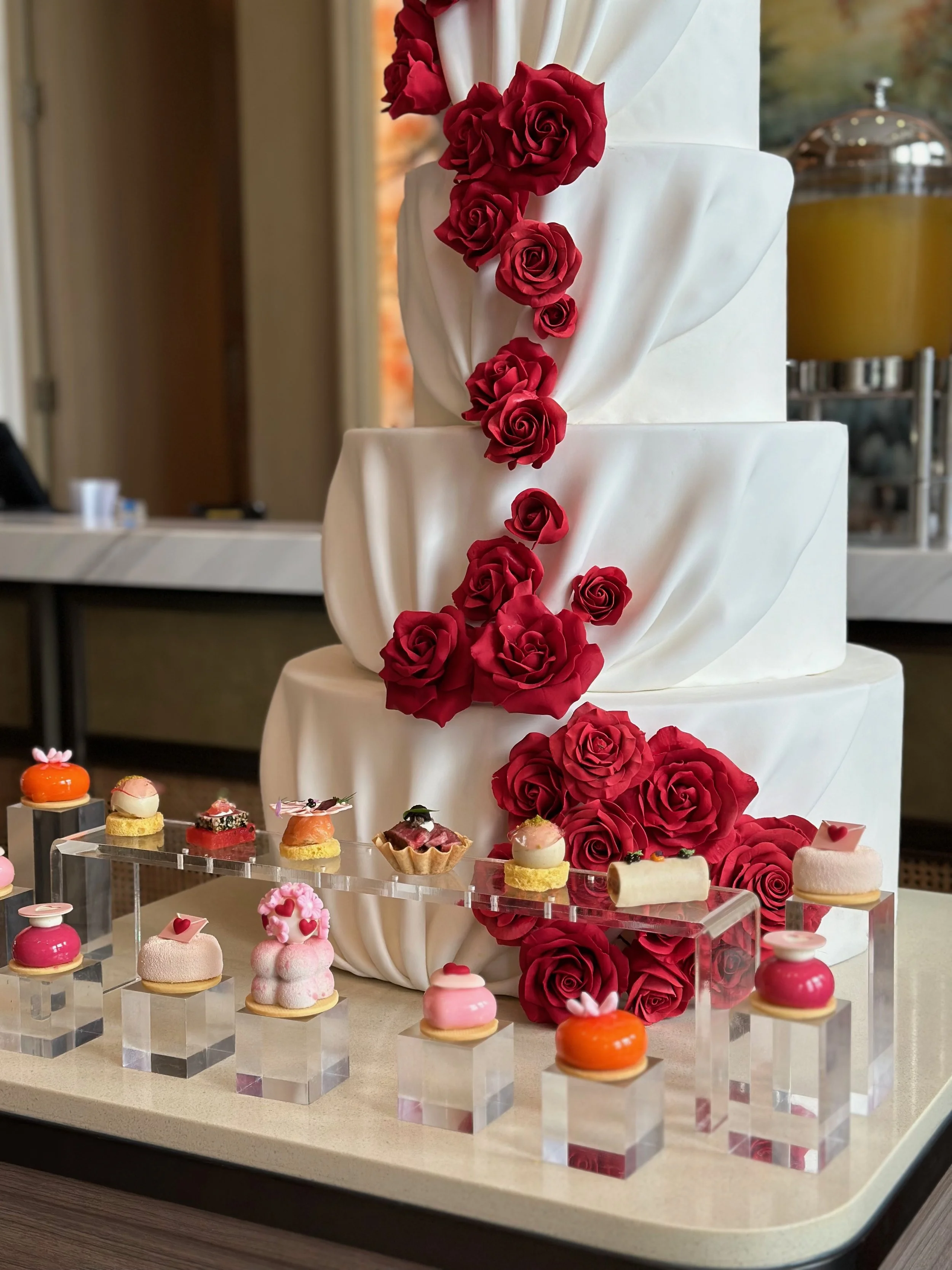 A three-tiered white cake decorated with red roses, with a display of assorted colorful petit fours in front.