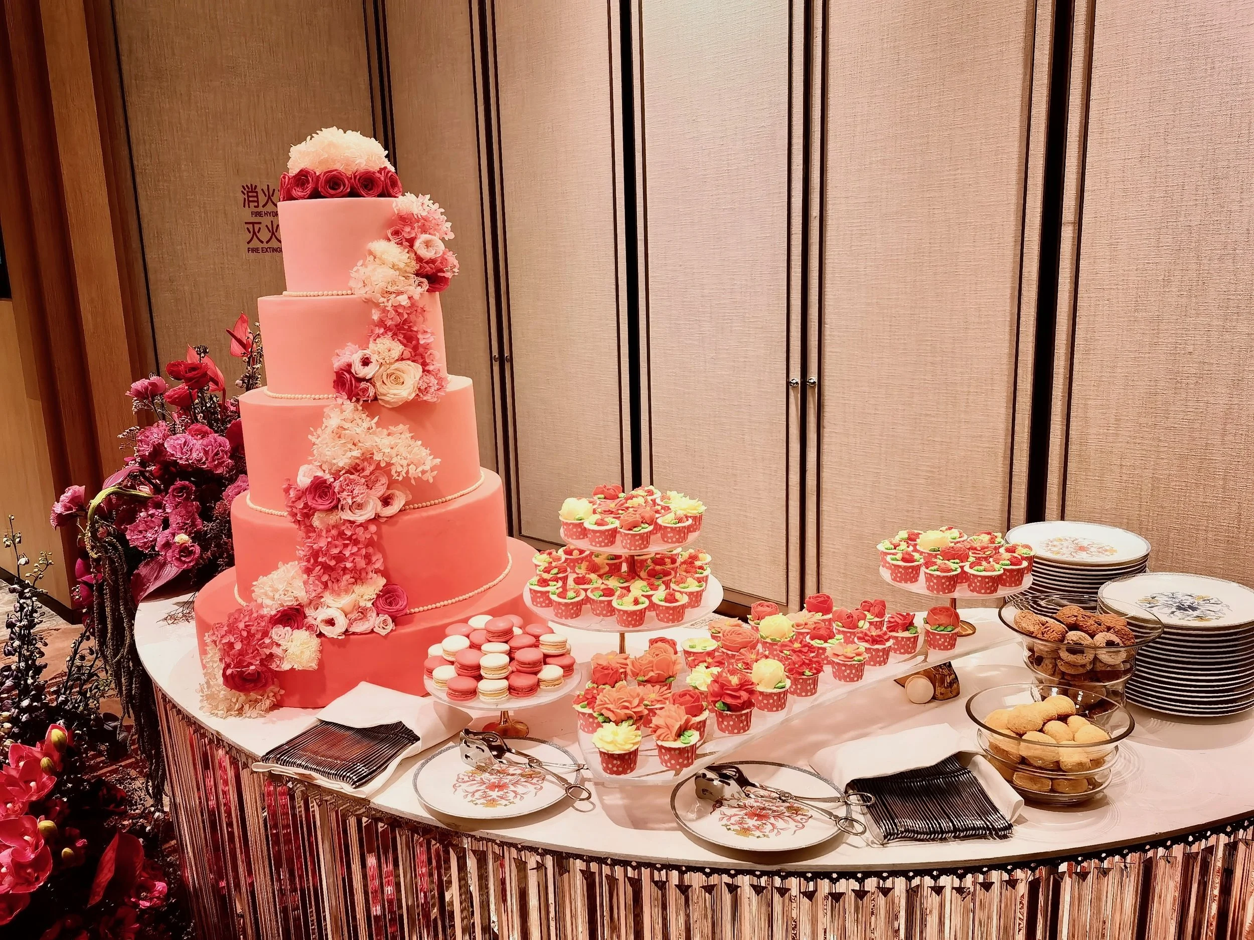 A pink, multi-tiered wedding cake decorated with pink and white flowers, surrounded by cupcakes with pink and yellow frosting, cookies, and plates on a table.
