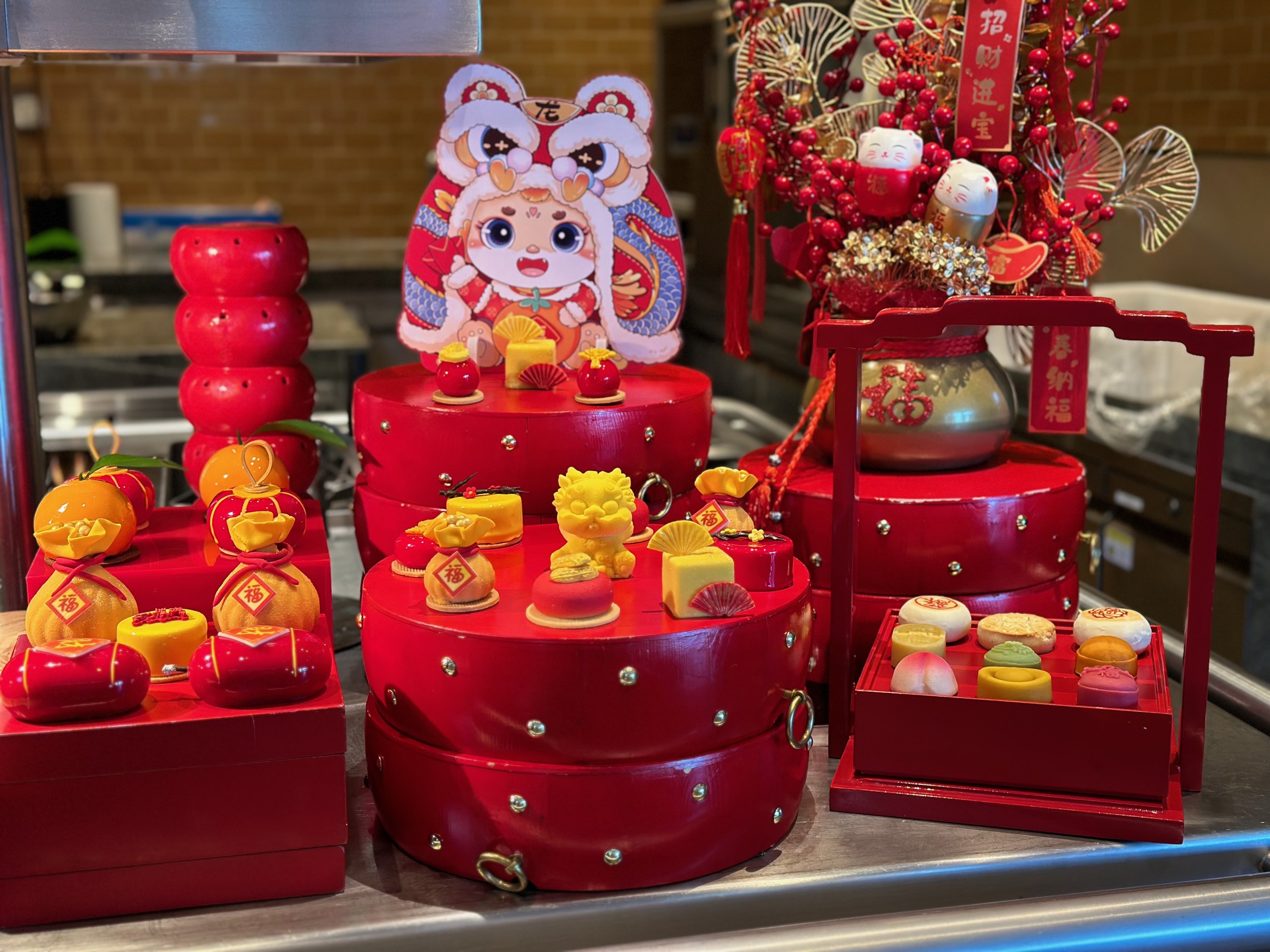 Chinese New Year decorations with red ornaments, flowers, and traditional symbols, including a cartoon figure, gold ingots, and snack boxes.