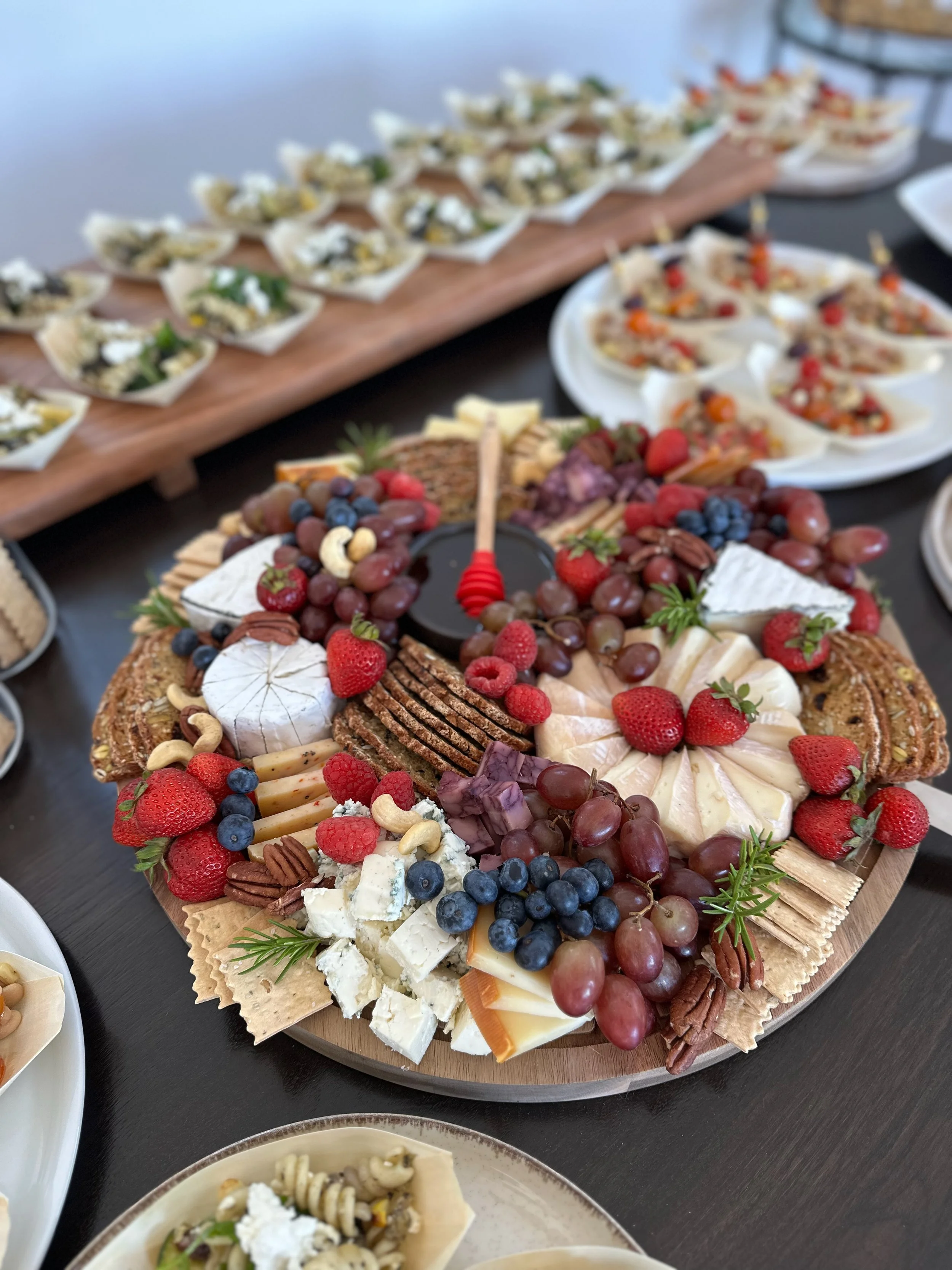 Charcuterie board with cheese, grapes, berries, crackers, and nuts. In the background, there are more appetizers on plates.