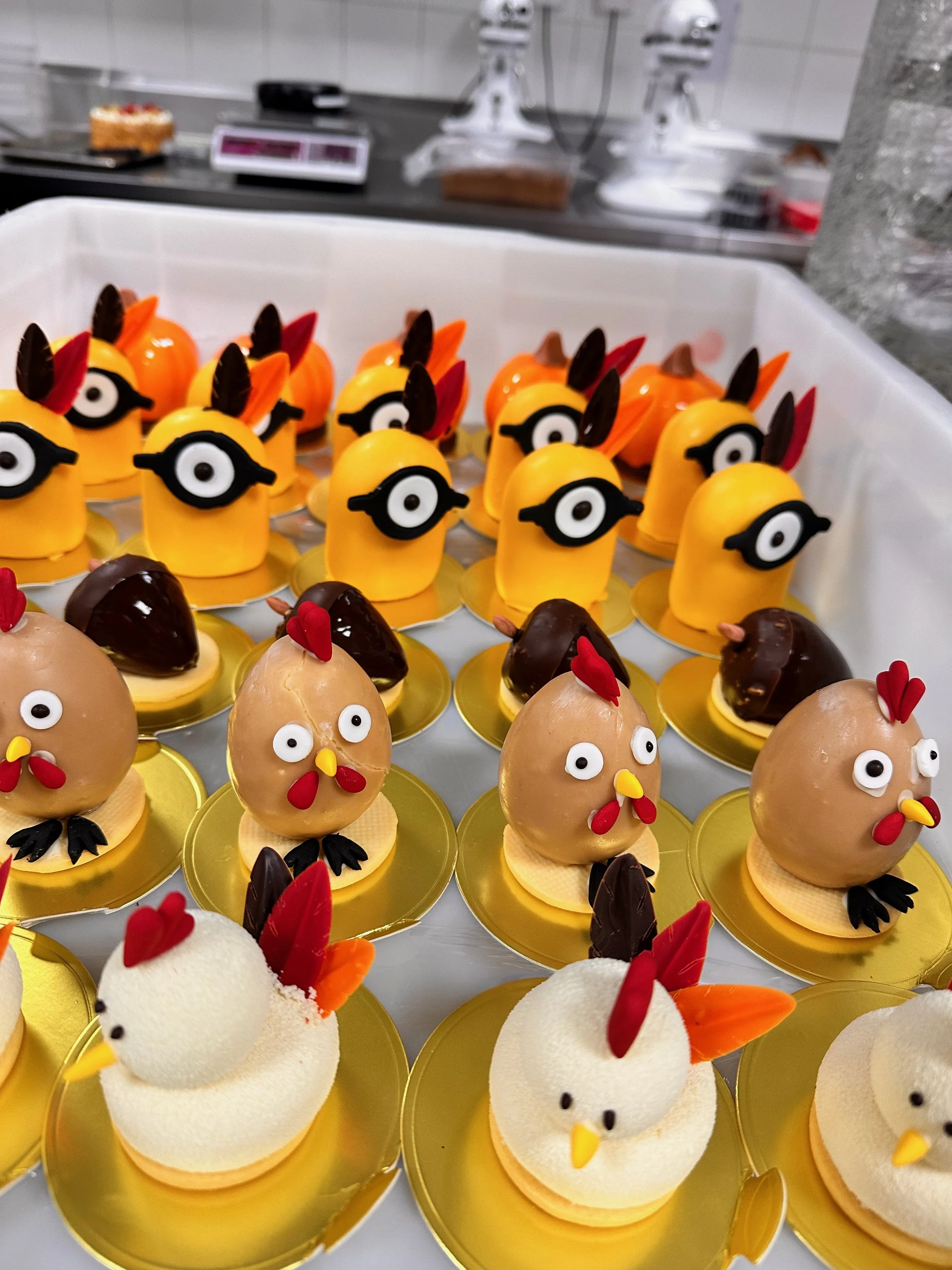 Decorative chicken and bird-themed desserts resembling chickens, chicks, and hens with colorful feathers and eyes on gold trays in a bakery or pastry shop.