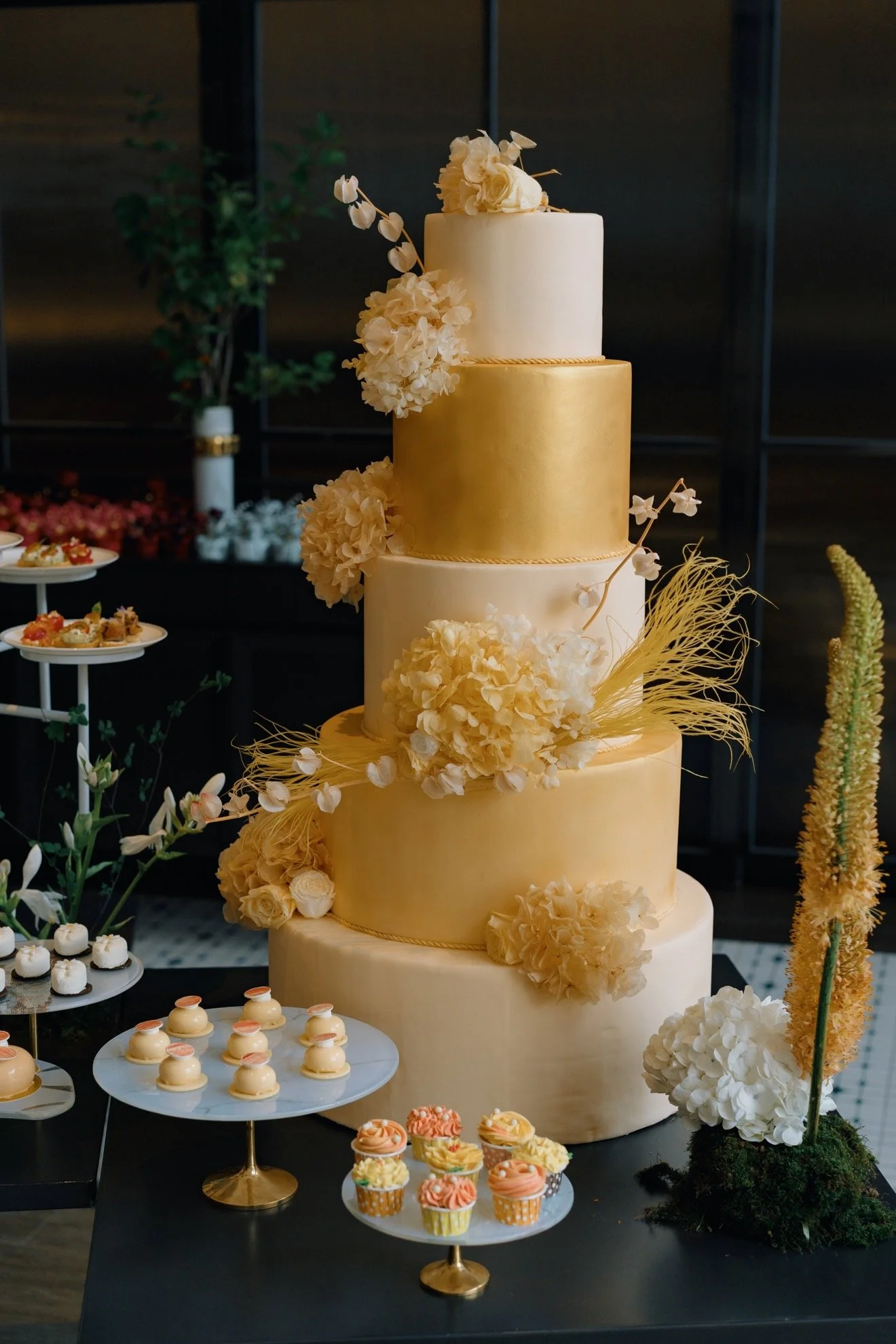 Four-tiered wedding cake decorated with white and gold fondant and floral accents, displayed with cupcakes and desserts on nearby platters.