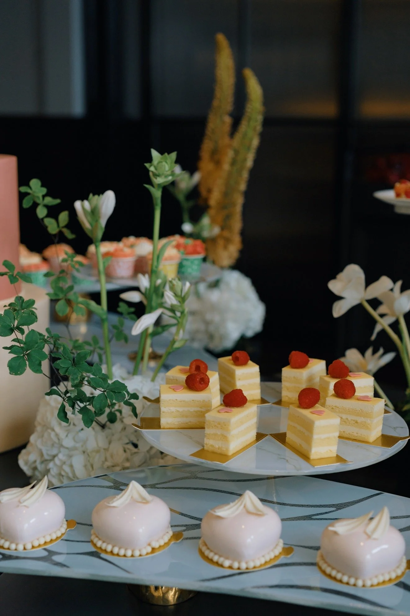 An elegant display of assorted desserts including layered cake squares topped with raspberries, and small heart-shaped cream pastries, arranged on a white platter with floral and greenery decorations in the background.