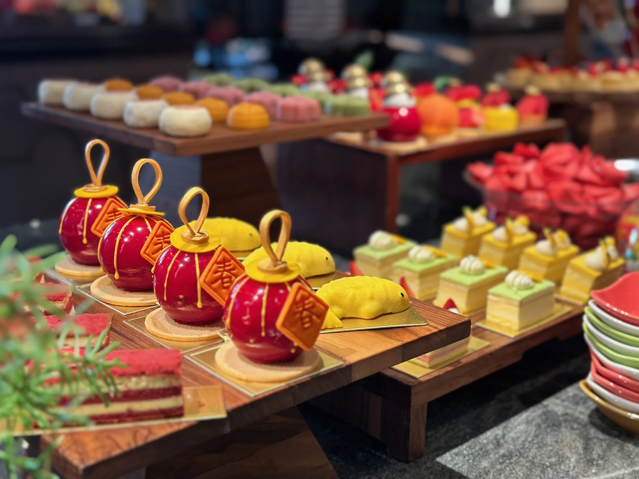 Assorted decorated pastries and desserts on display at a bakery or dessert shop, including red themed pastries with gold accents, yellow plush-like desserts, layered cakes with green frosting, and various other colorful confections.
