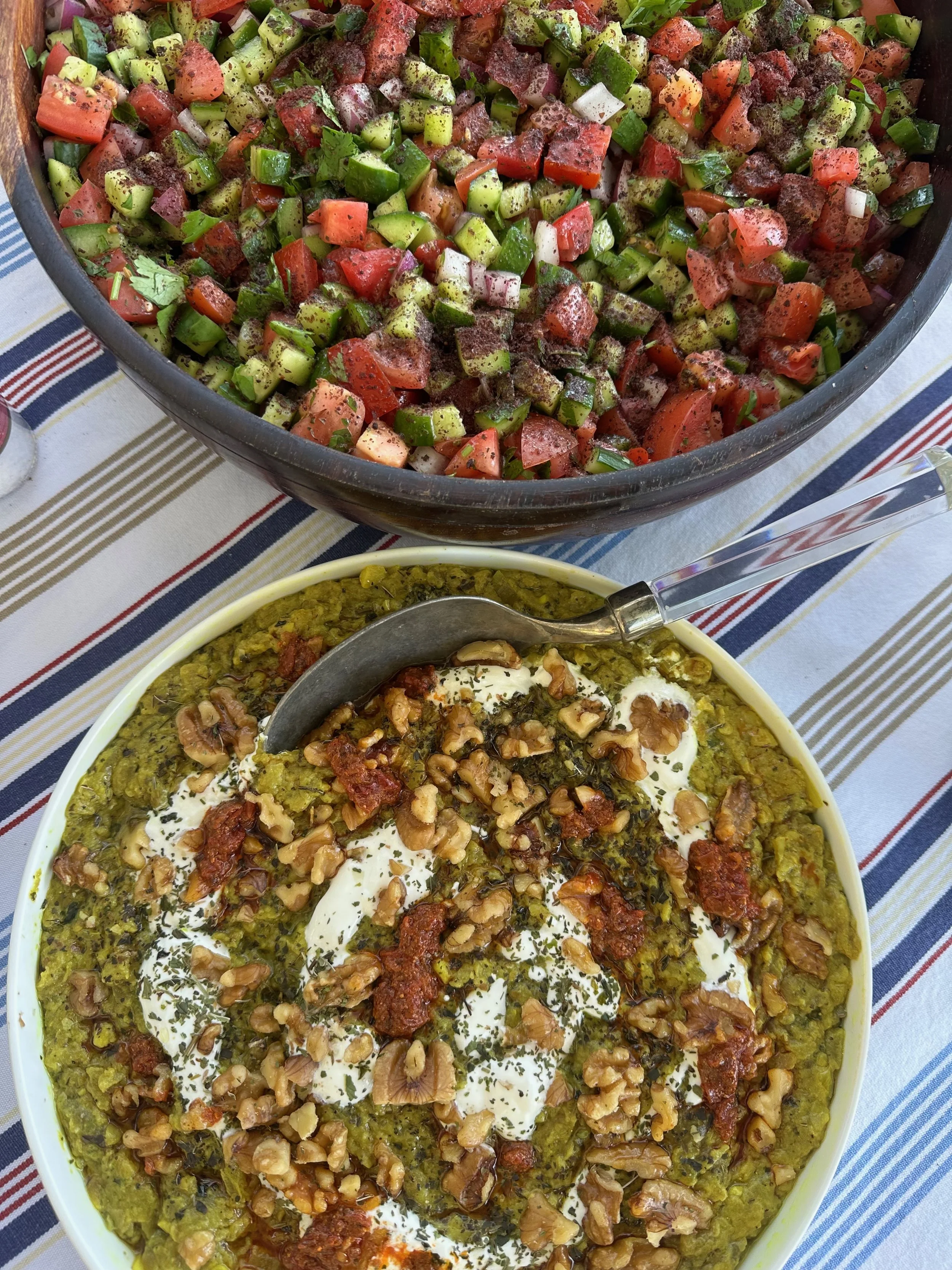 A bowl of cooked green chili topped with sour cream, chopped walnuts, herbs, and some red sauce, and a large bowl of chopped salad with tomatoes, cucumber, and green onions on a striped tablecloth.