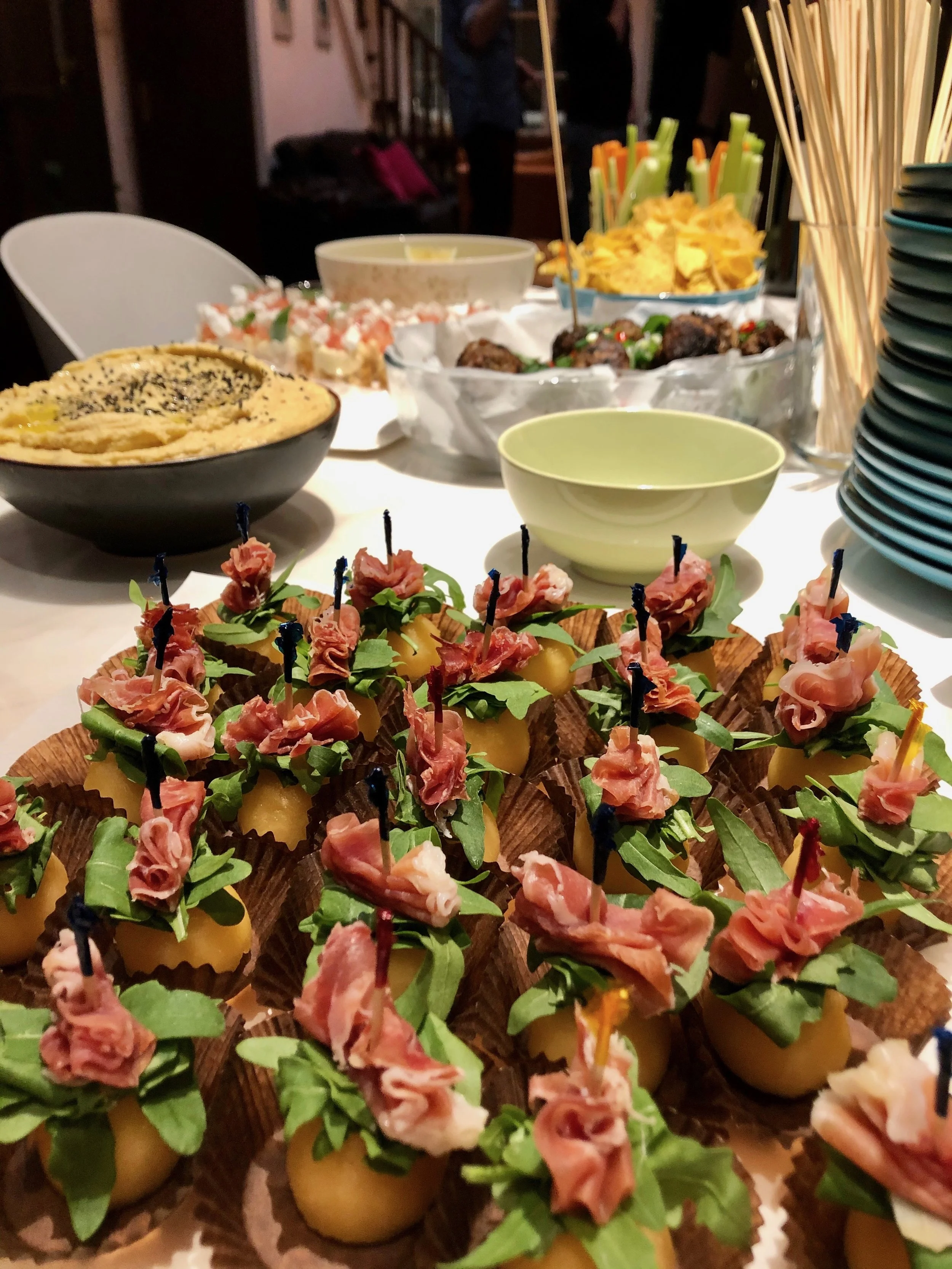 Appetizers with cured meats, cheese, and greens on a festive buffet table with other dishes in the background.