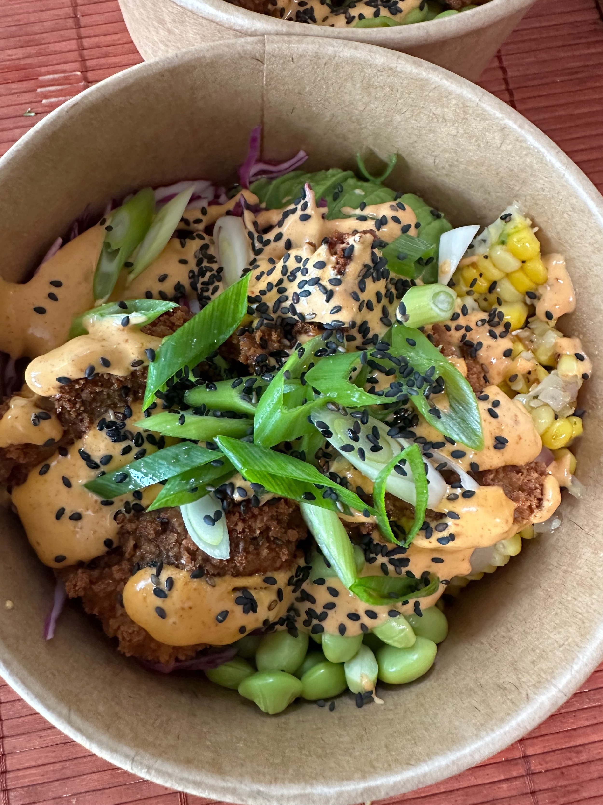 A bowl of Asian-inspired food with fried meat, green onions, edamame, corn, sesame seeds, and spicy sauce.