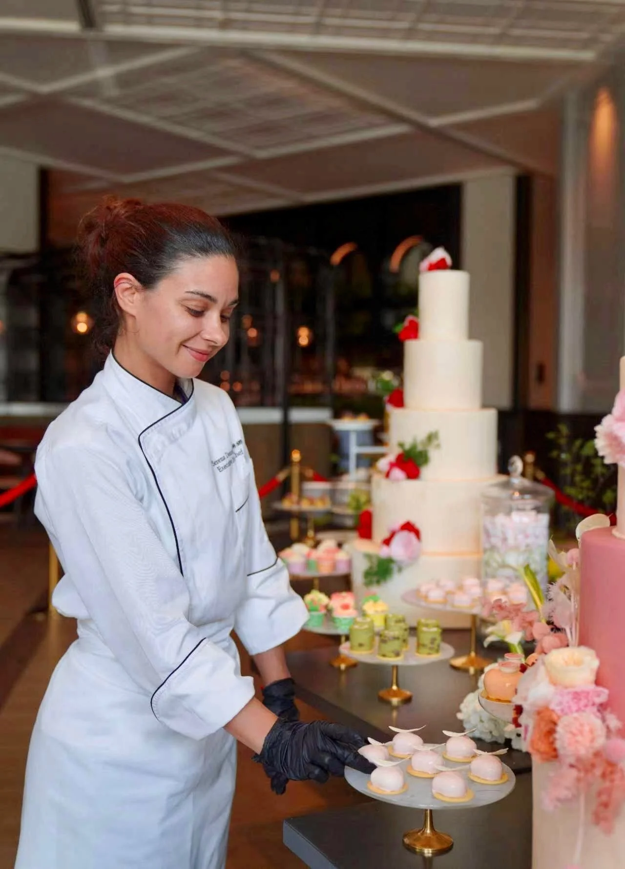 A woman in a white chef's coat and black gloves is preparing pastries at a dessert table with a wedding cake and other sweets in the background.