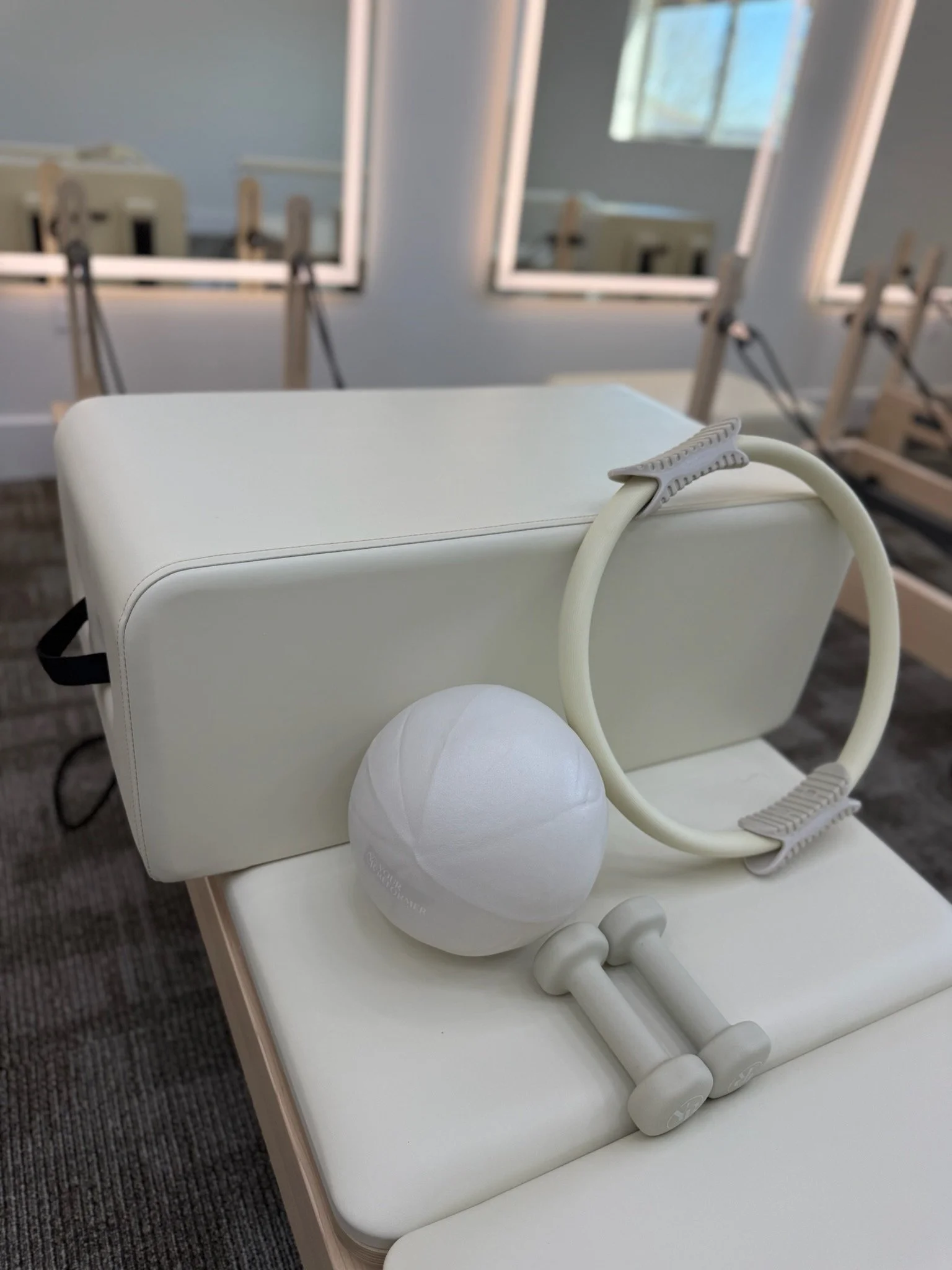 Medical equipment including a white deflated exercise ball, a white textured resistance ring, and a small pair of white dumbbells placed on a white treatment table in a therapy or gym room with windows and mirrors.