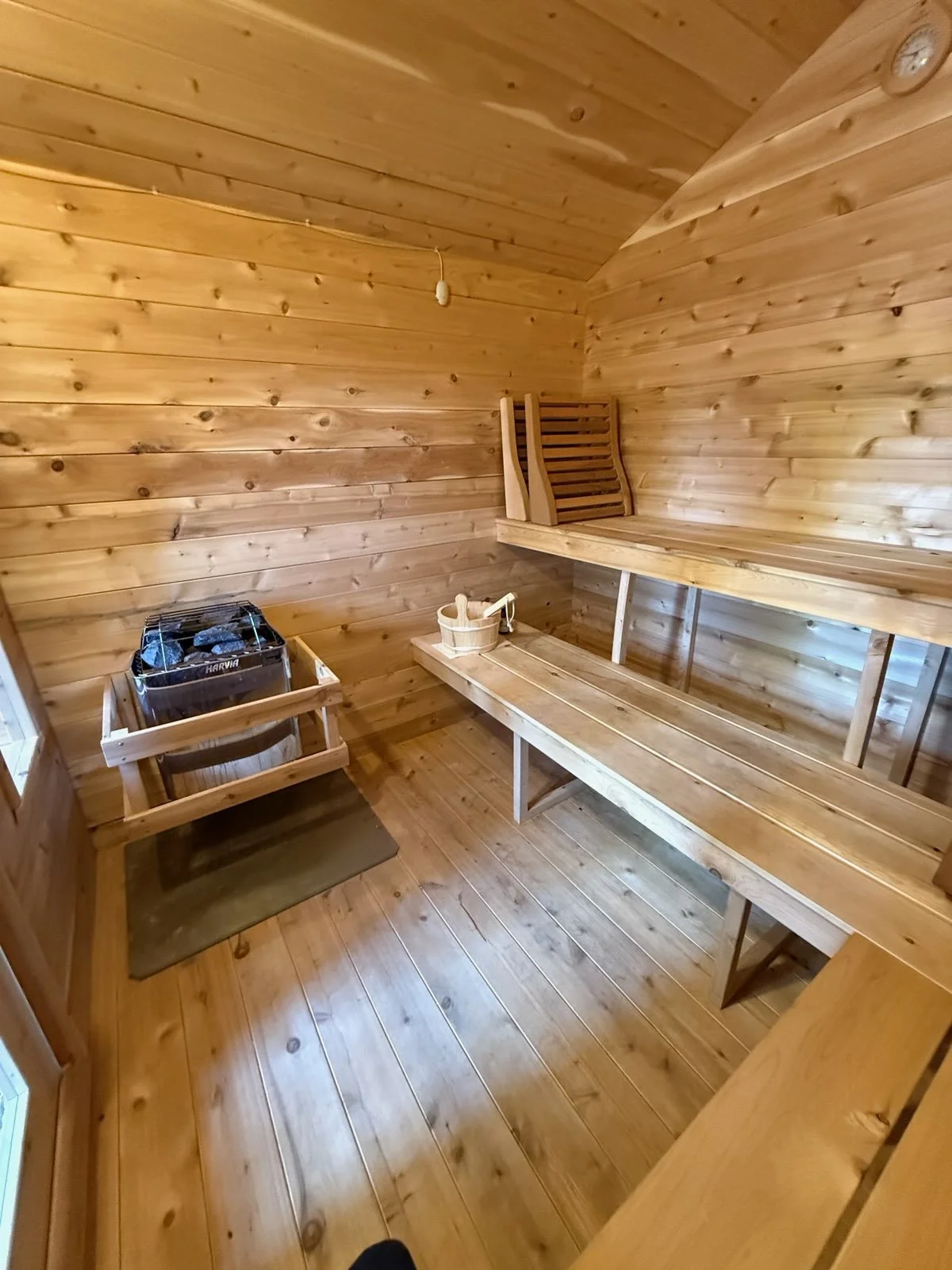 A small wooden sauna room with a sauna heater with stones, a wooden bucket, and a ladle, along with wooden benches and a wall-mounted chair, all made of light-colored wood.