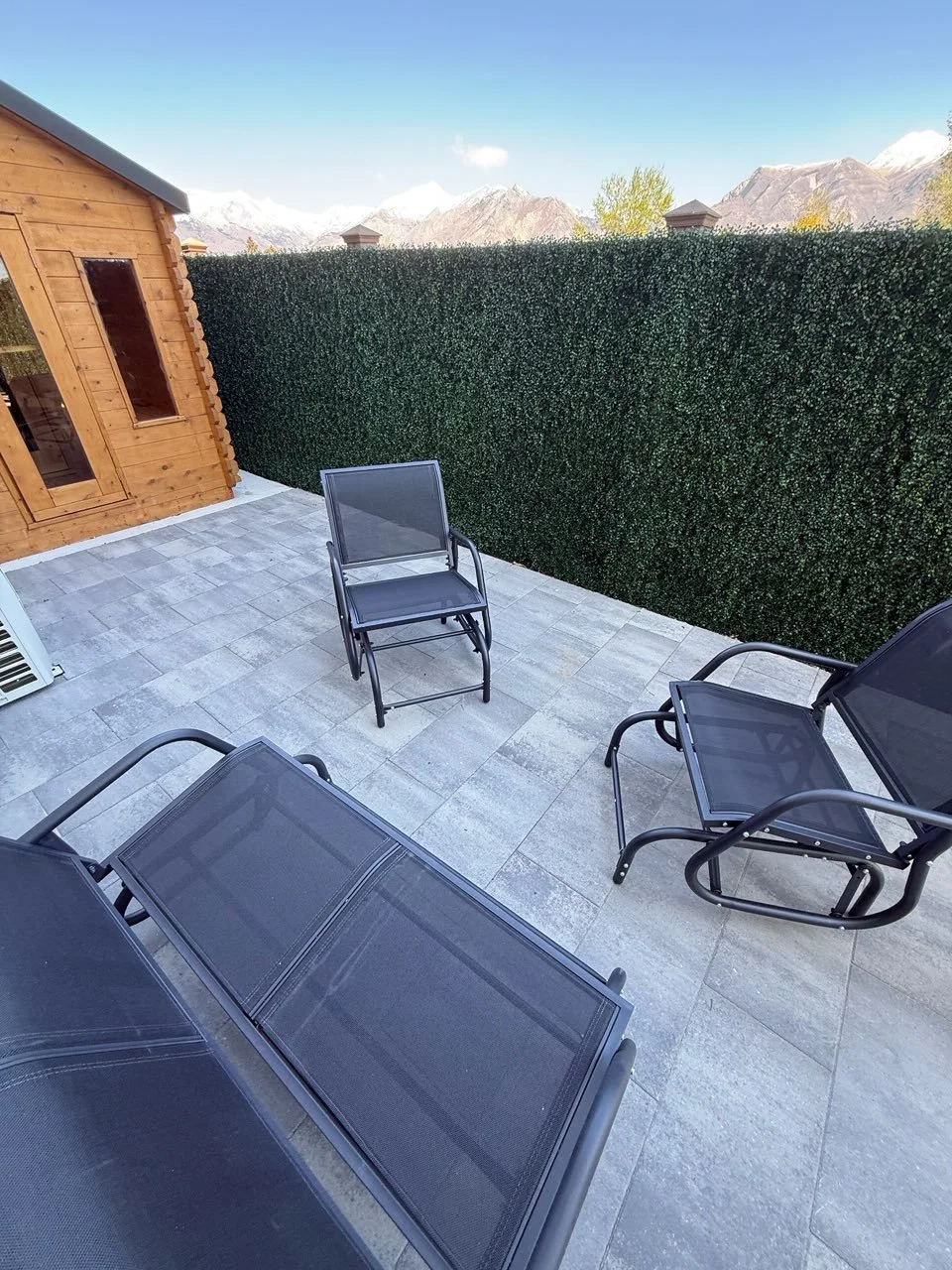 Outdoor patio with black mesh lounge chair, single black mesh dining chair, black mesh armchair, on grey stone tiles, backed by a green hedge, with mountains and blue sky in the background.