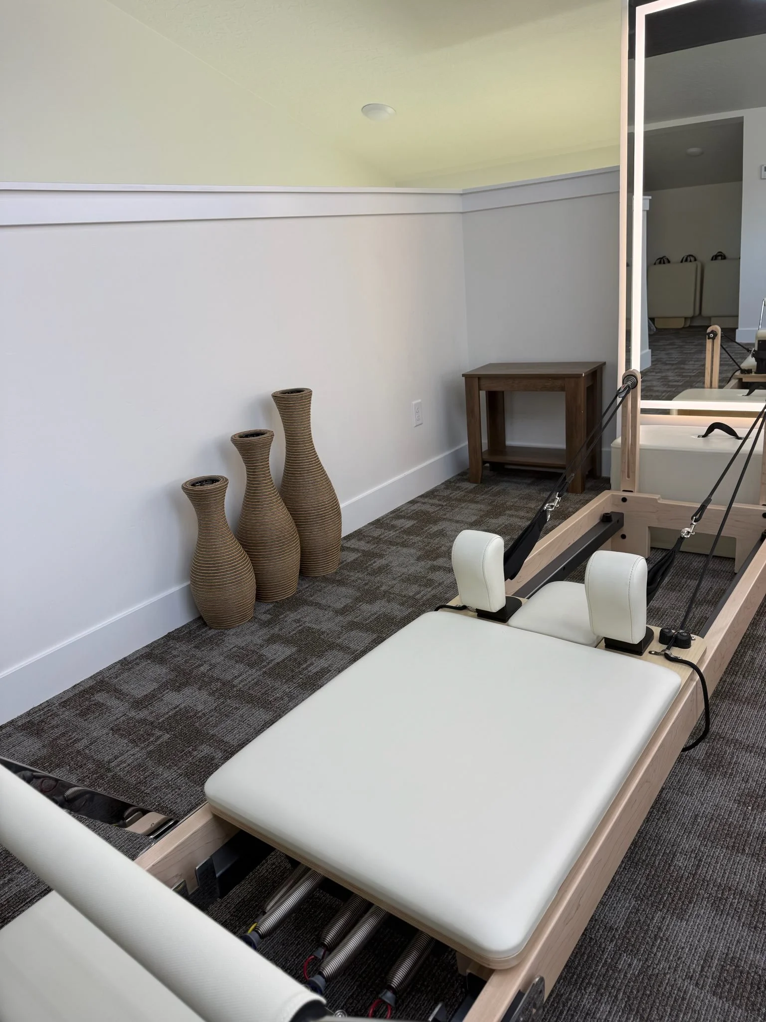 A medical or therapy table with padded surface and adjustable headrest, located in a room with white walls, a small wooden side table, decorative vases, and a mirror.