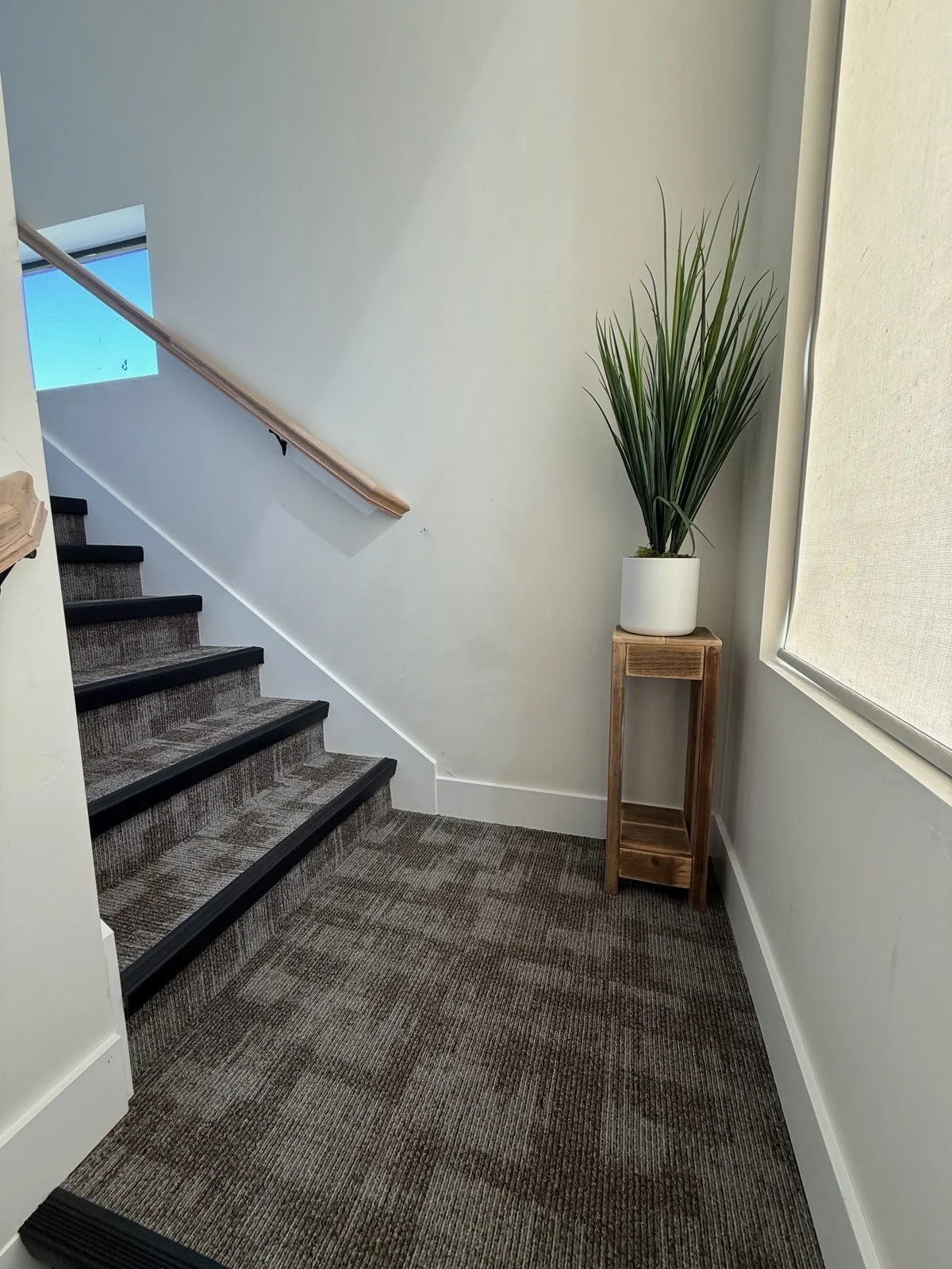 Interior view of a staircase with carpeted steps, a wooden handrail, a potted plant on a wooden stand, and a window with a light-colored shade.