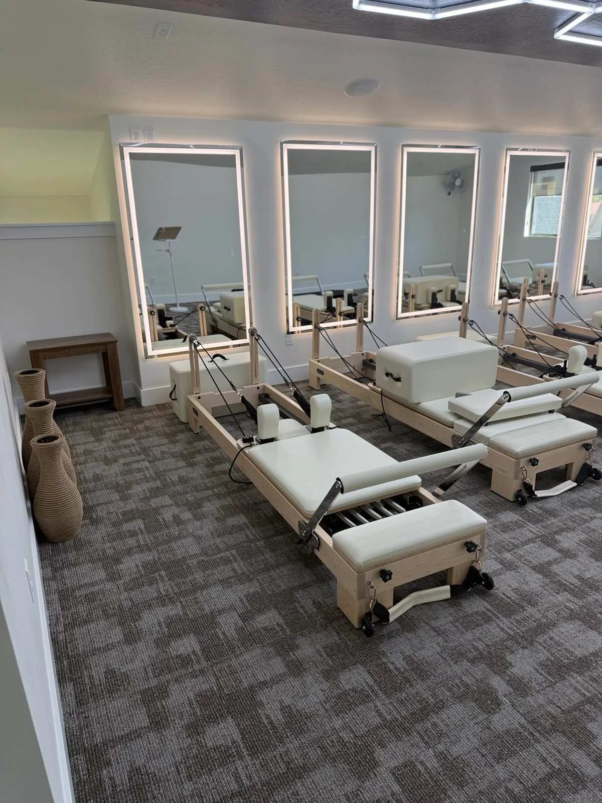 A room with three Pilates reformer machines, mirrors on the wall, and a window with blinds, decorated in neutral tones.