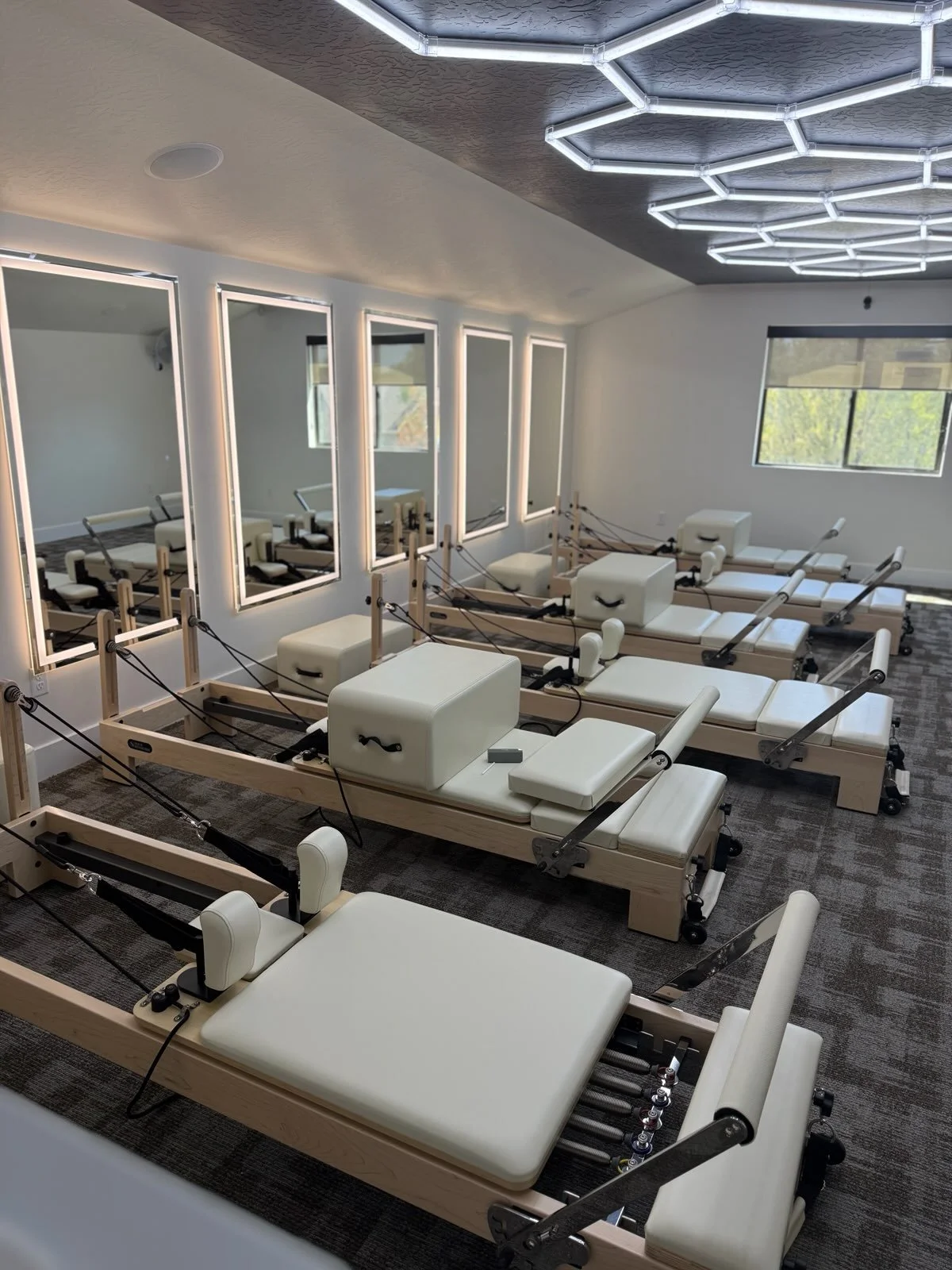 A room with multiple Pilates reformer machines with white padded surfaces, facing a wall of mirrors and windows with daylight outside. The ceiling has geometric LED lights.