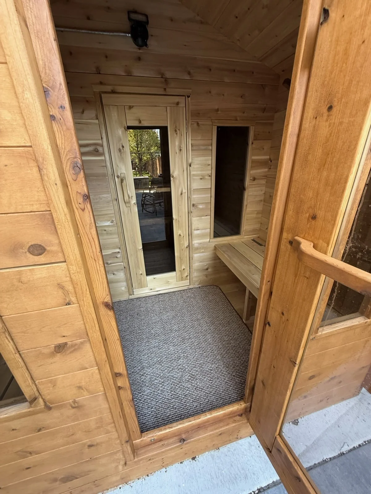 A small wooden porch with a black doormat, a glass door, and a window. Inside is a bench and wooden walls.