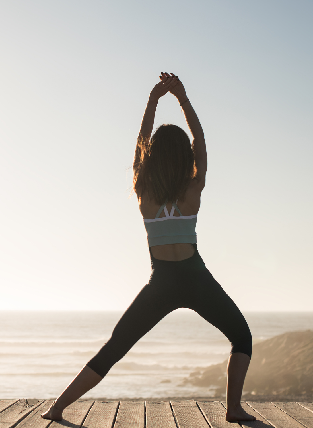 A woman practicing yoga outdoors on a wooden platform by the sea at sunset, stretching with arms overhead and legs apart.