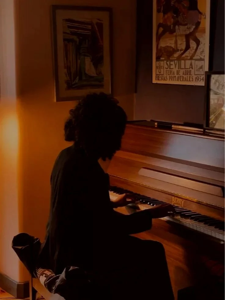 Person with curly hair playing a wooden upright piano in a dimly lit room with wall art and a large window.