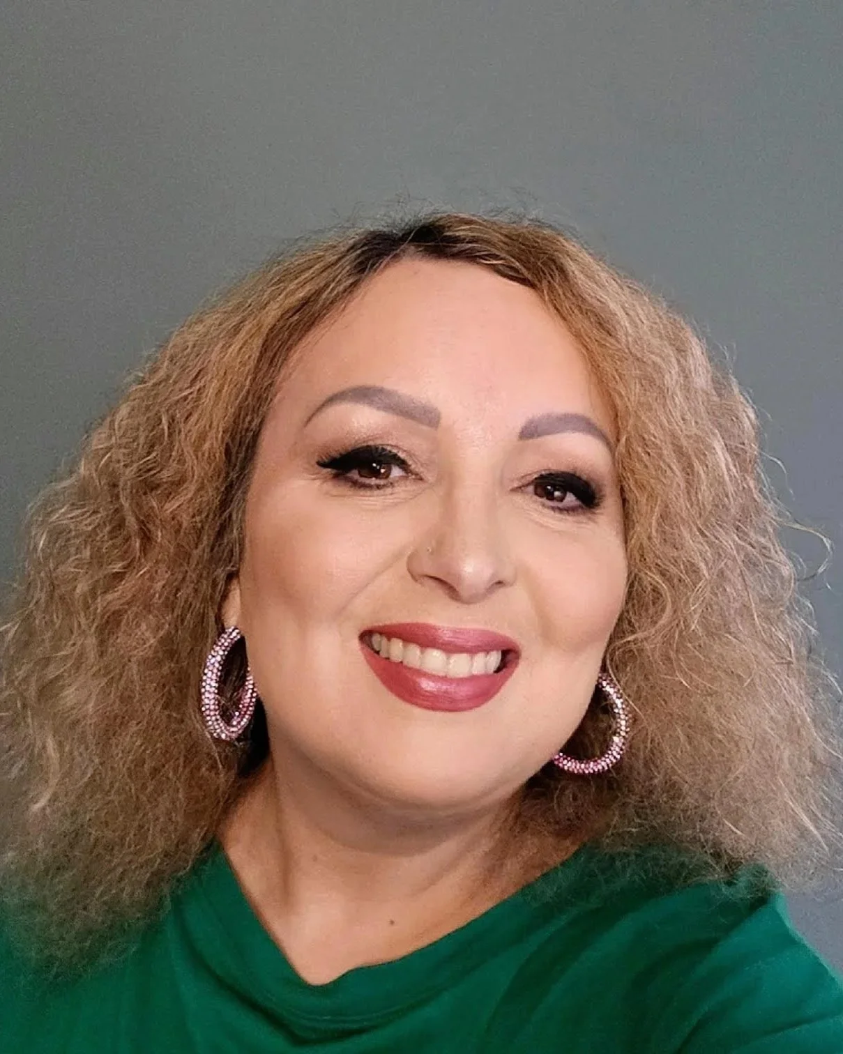 A woman with curly hair, wearing pink earrings and a green top, smiling at the camera.