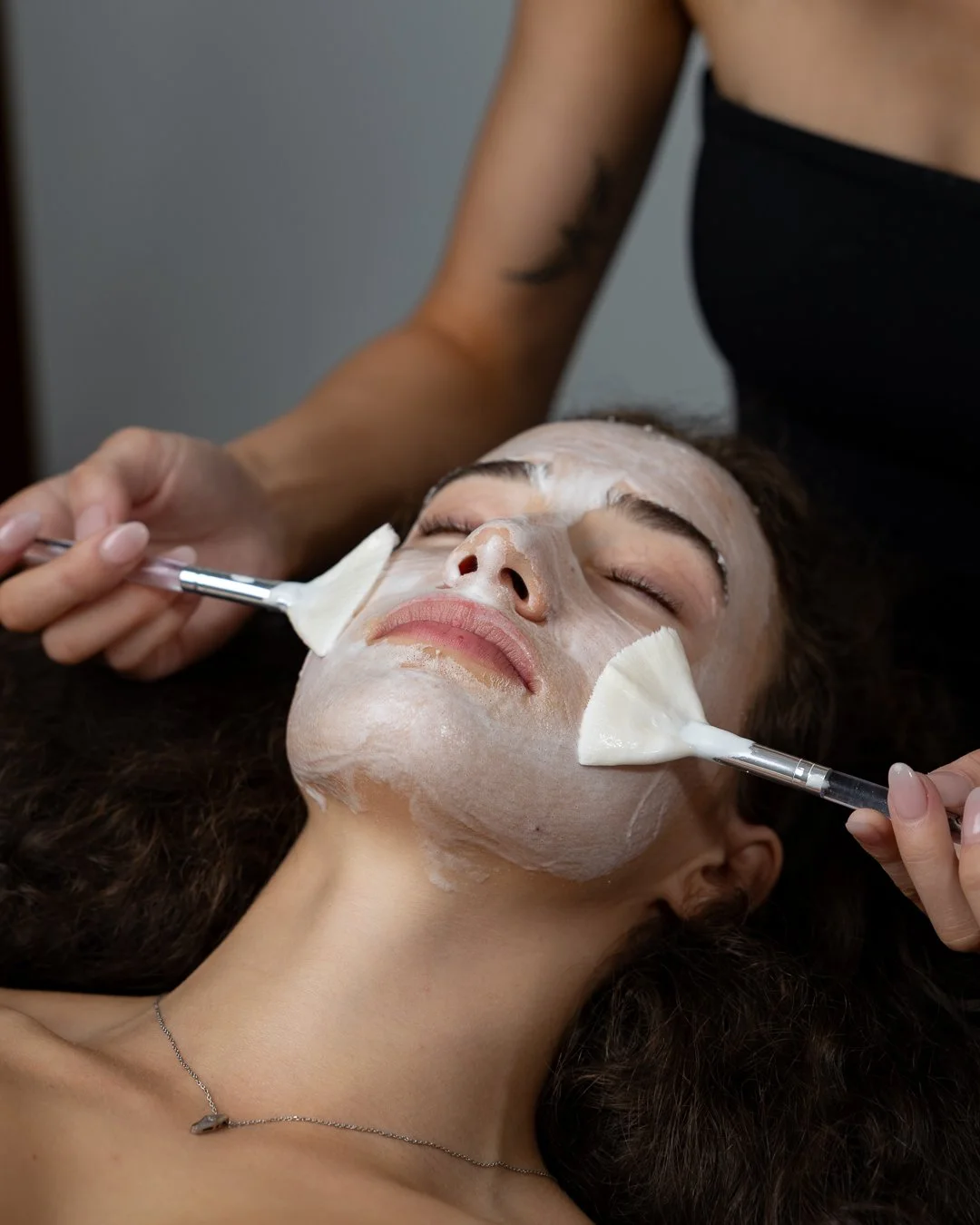 A woman lying down with facial cream mask being applied using brushes during a skincare treatment.