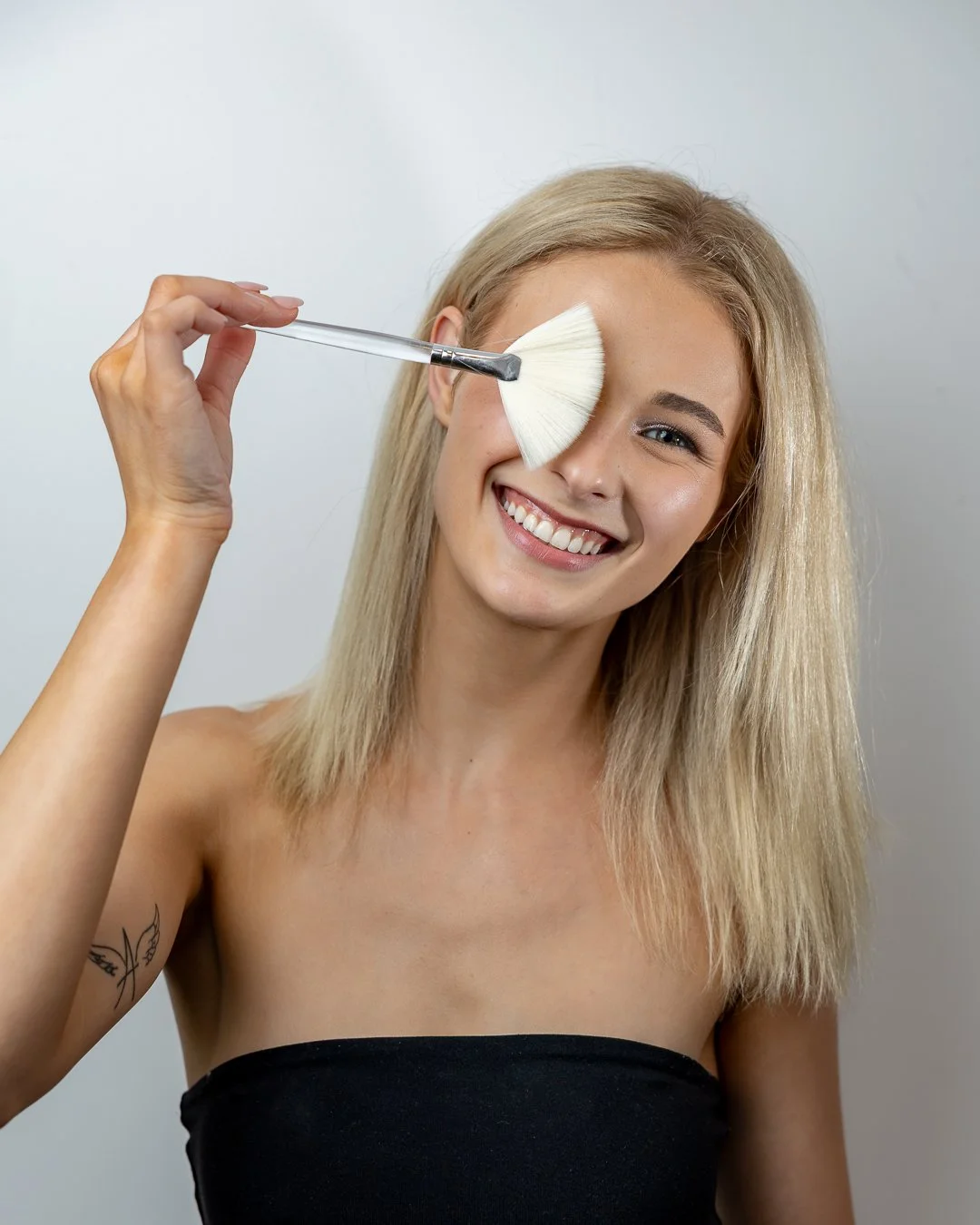 A woman with blonde hair smiling as she holds a fan-shaped makeup brush to her eye, wearing a black strapless top.