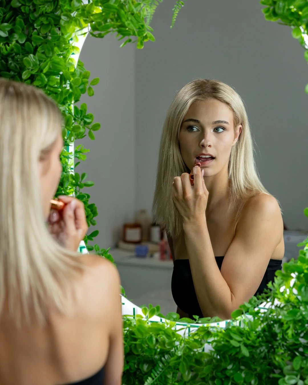 A young woman with long blonde hair applying lipstick in front of a mirror surrounded by green plants.
