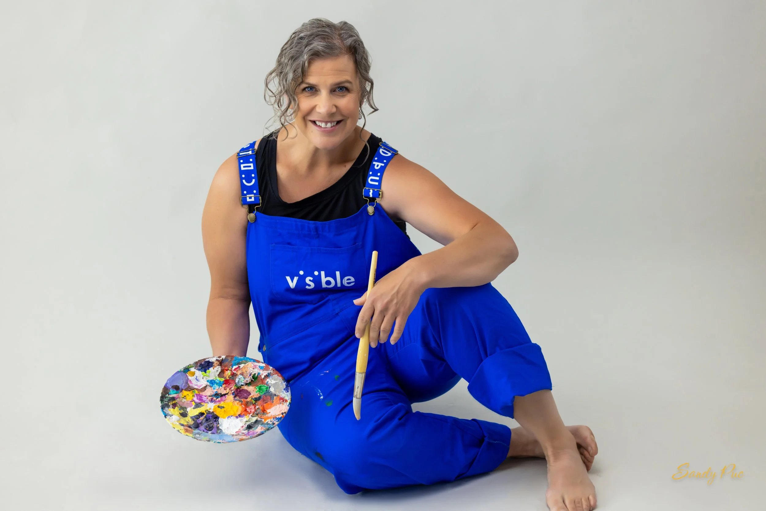 A woman with curly gray hair, smiling, wearing a black top and blue overalls with 'visible' printed on the front. She is sitting on the floor, holding a paint palette with colorful paints in her left hand and a paintbrush in her right hand. The background is plain white, and the name 'Sandy Puc' appears in the bottom right corner.