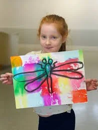 Young girl with red hair holding colorful painting of a dragonfly with purple, blue, and red wings on a bright background.