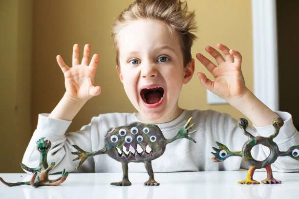 Child with surprised expression sitting at a table, with three colorful monster-shaped clay sculptures in front of him.