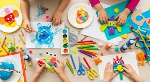 Children's hands engage in arts and crafts with plates holding colorful paint and craft supplies like scissors, markers, and glue, surrounded by paper and decorated craft projects.