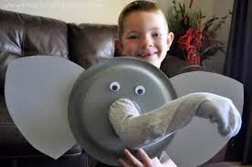 Child smiling while holding a homemade elephant craft with a paper plate face and large paper ears.