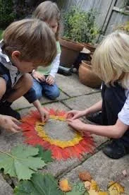 Three children gathered around a colorful sand art project on the ground outdoors, working together on a flower-shaped design with fall leaves and small pumpkins nearby.