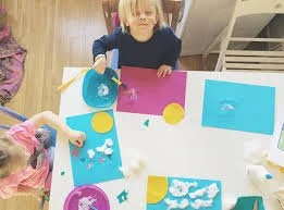 Children engaging in arts and crafts, working with colorful paper, cotton balls, and craft supplies at a table.