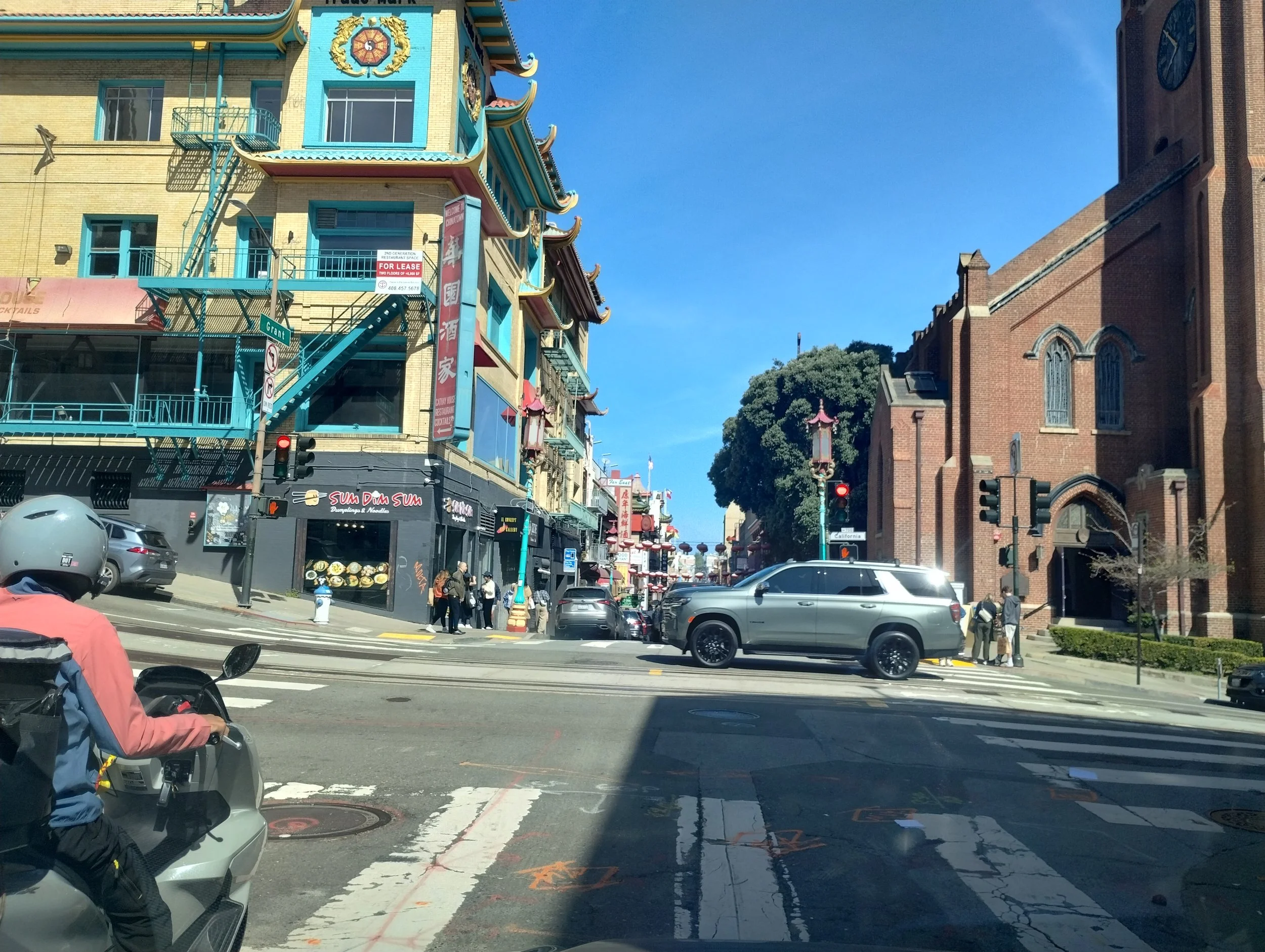 Street scene in Chinatown with colorful buildings featuring Asian architectural elements, people walking on the sidewalk, cars parked along the street, and a clear blue sky overhead.