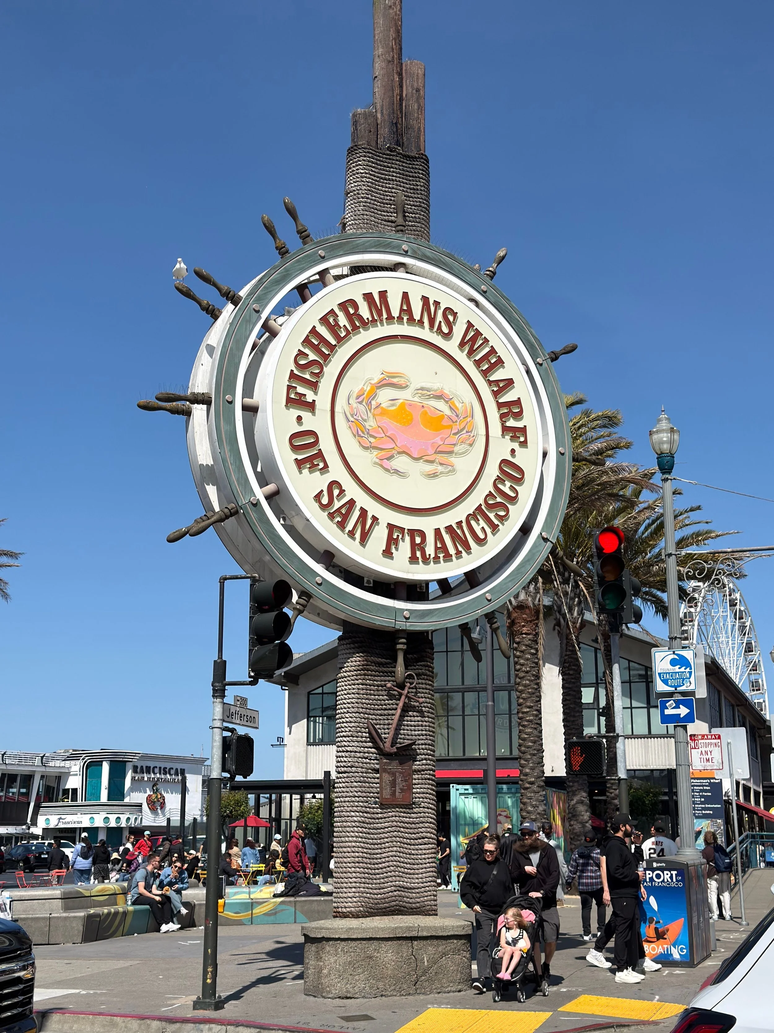 A busy city scene at Fisherman's Wharf in San Francisco, featuring a large nautical-themed clock with a crab illustration, people walking, sitting, and shopping, palm trees, street signs, and a bright blue sky.