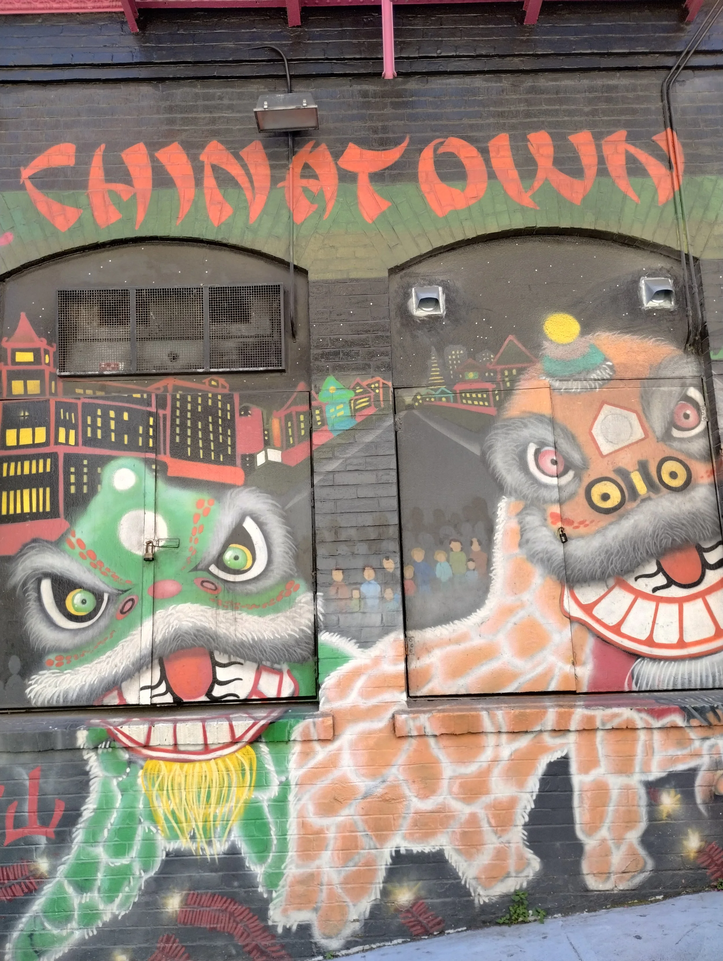 Colorful murals on a brick wall depicting a traditional Chinese lion dance with a green lion on the left and an orange lion on the right, along with cityscape and mountain scenery in the background.