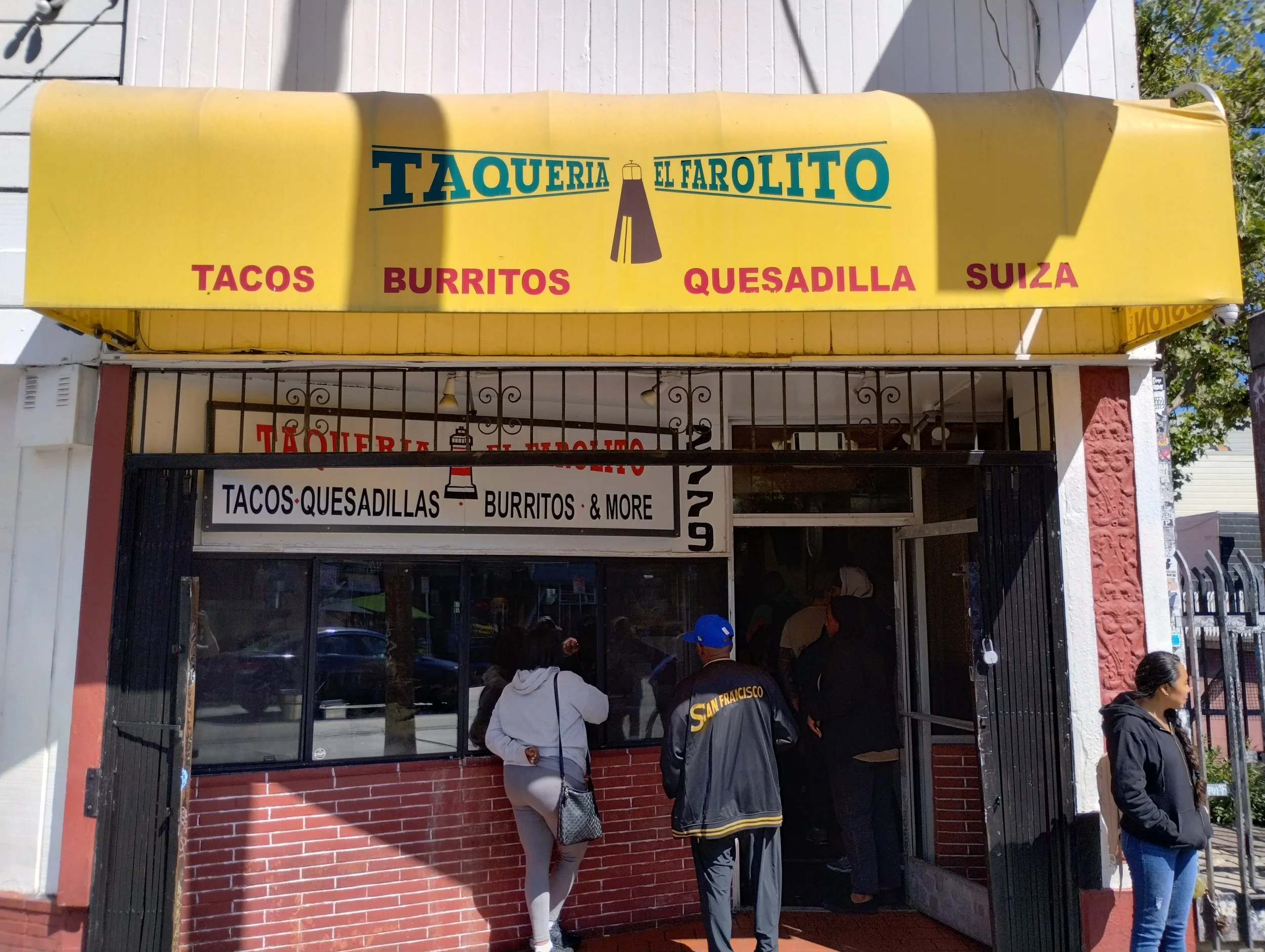 People standing in line outside Taqueria El Farolito restaurant with a yellow signboard displaying menu items like tacos, burritos, quesadillas, and Swiss.