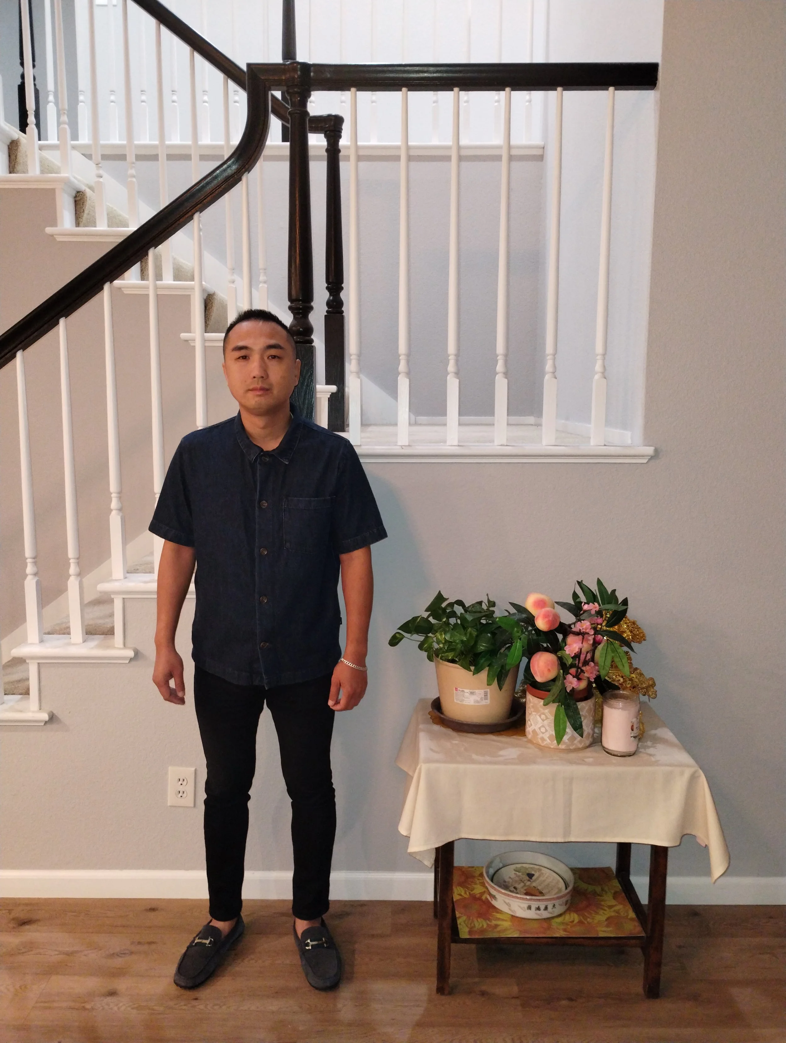 A young man with short dark hair, wearing a dark short-sleeve button-up shirt, black pants, and black slip-on shoes, standing indoors next to a table with potted plants and decorative items, in front of a staircase with white railings and a wall with vertical white paneling.
