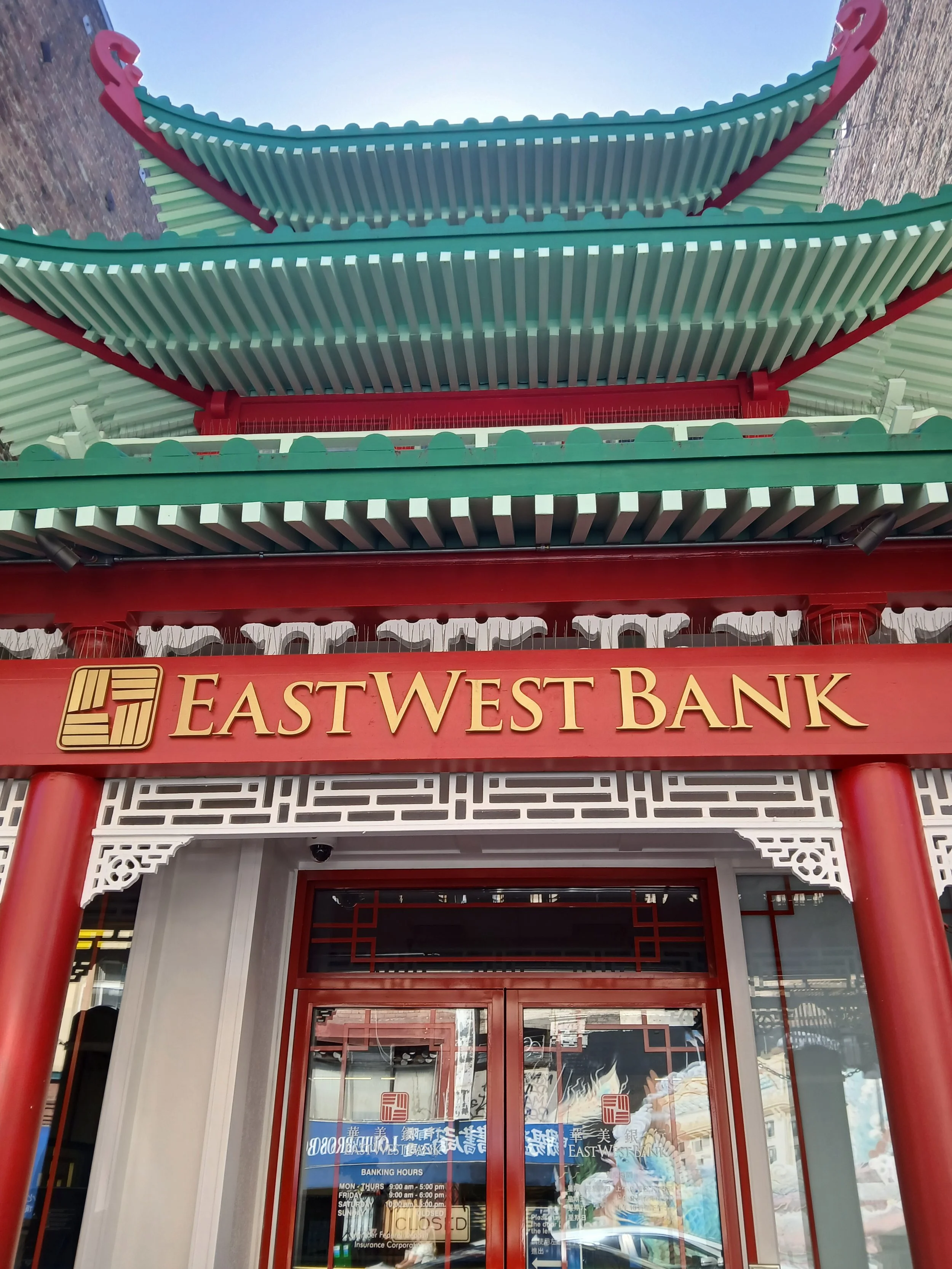 The entrance of East West Bank featuring traditional Chinese architectural design with red columns, decorative white lattice work, and a roof with teal and red accents.