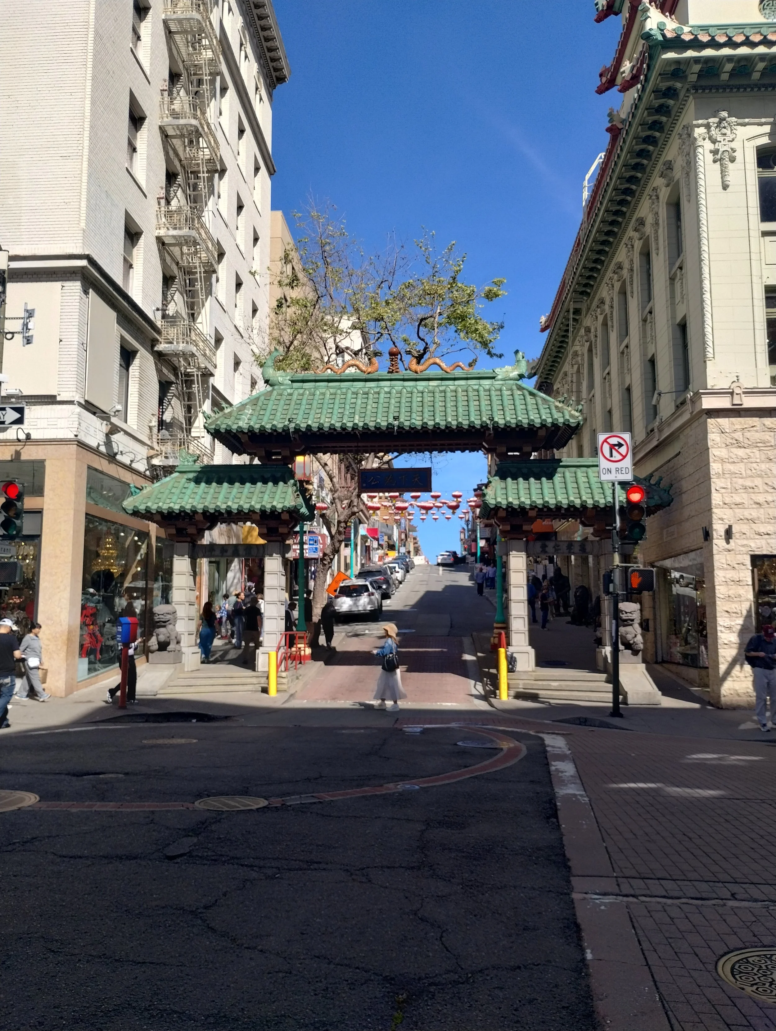 Street scene with a traditional Chinese-style gate in the foreground, buildings on either side, and a clear blue sky above. Pedestrians walk on the sidewalk, and a traffic light is visible.